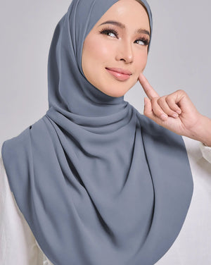 A woman wearing a draped dusty blue-gray hijab and a white blouse, posing against a light gray studio background.