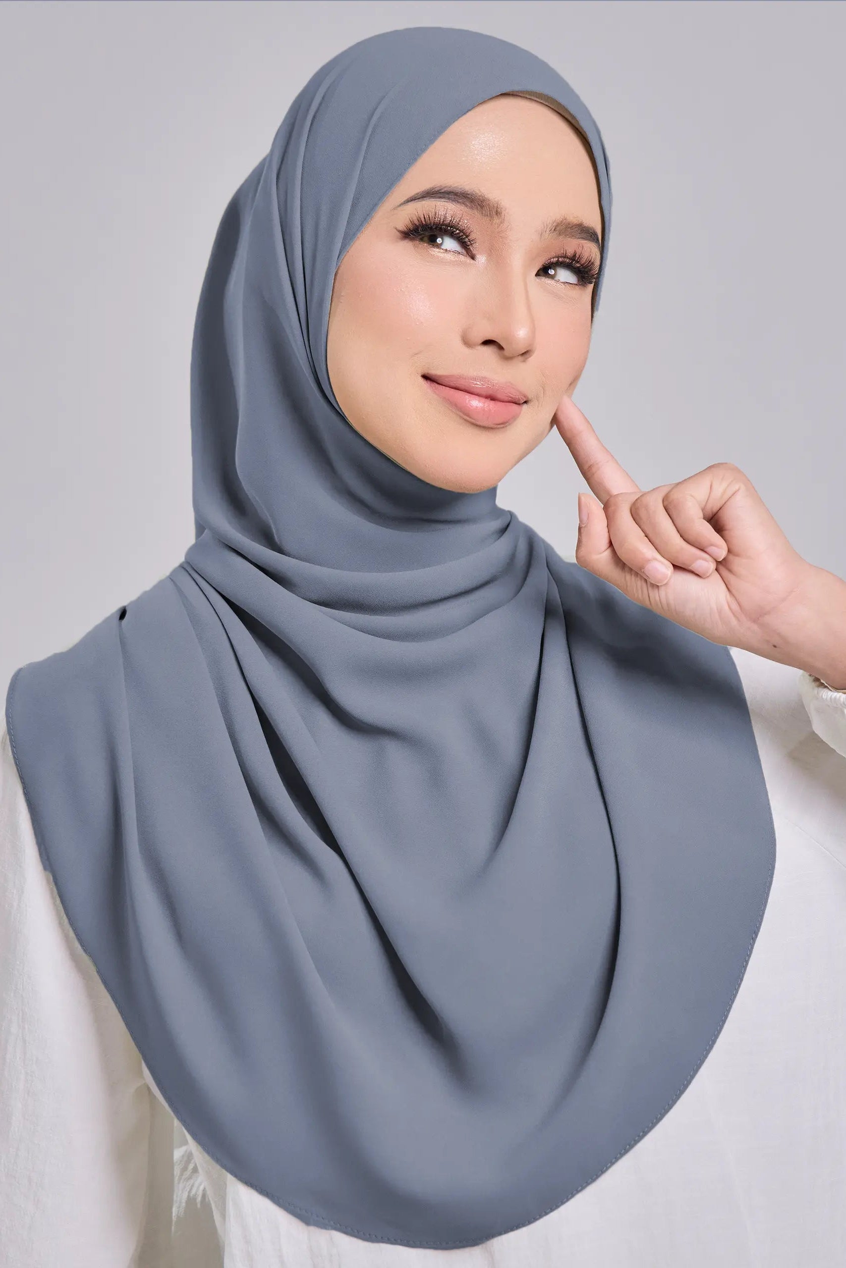 A woman wearing a draped dusty blue-gray hijab and a white blouse, posing against a light gray studio background.