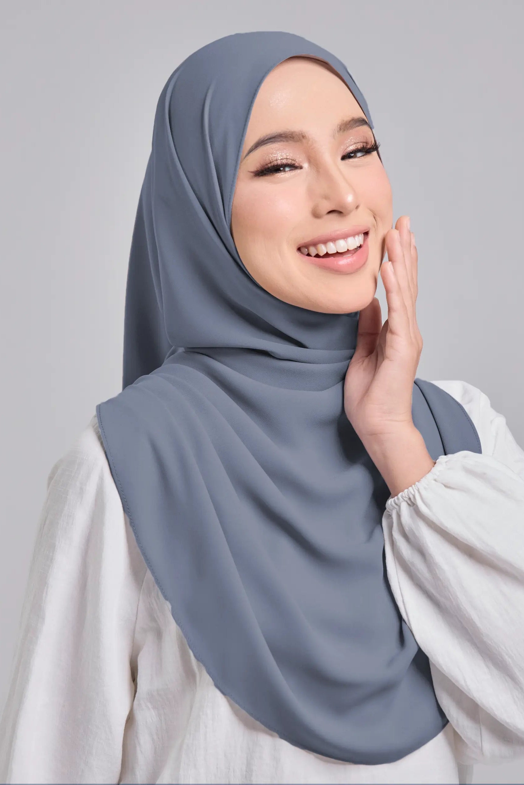 A woman wearing a slate blue-gray hijab and a white long-sleeve blouse, posing against a solid light gray background.