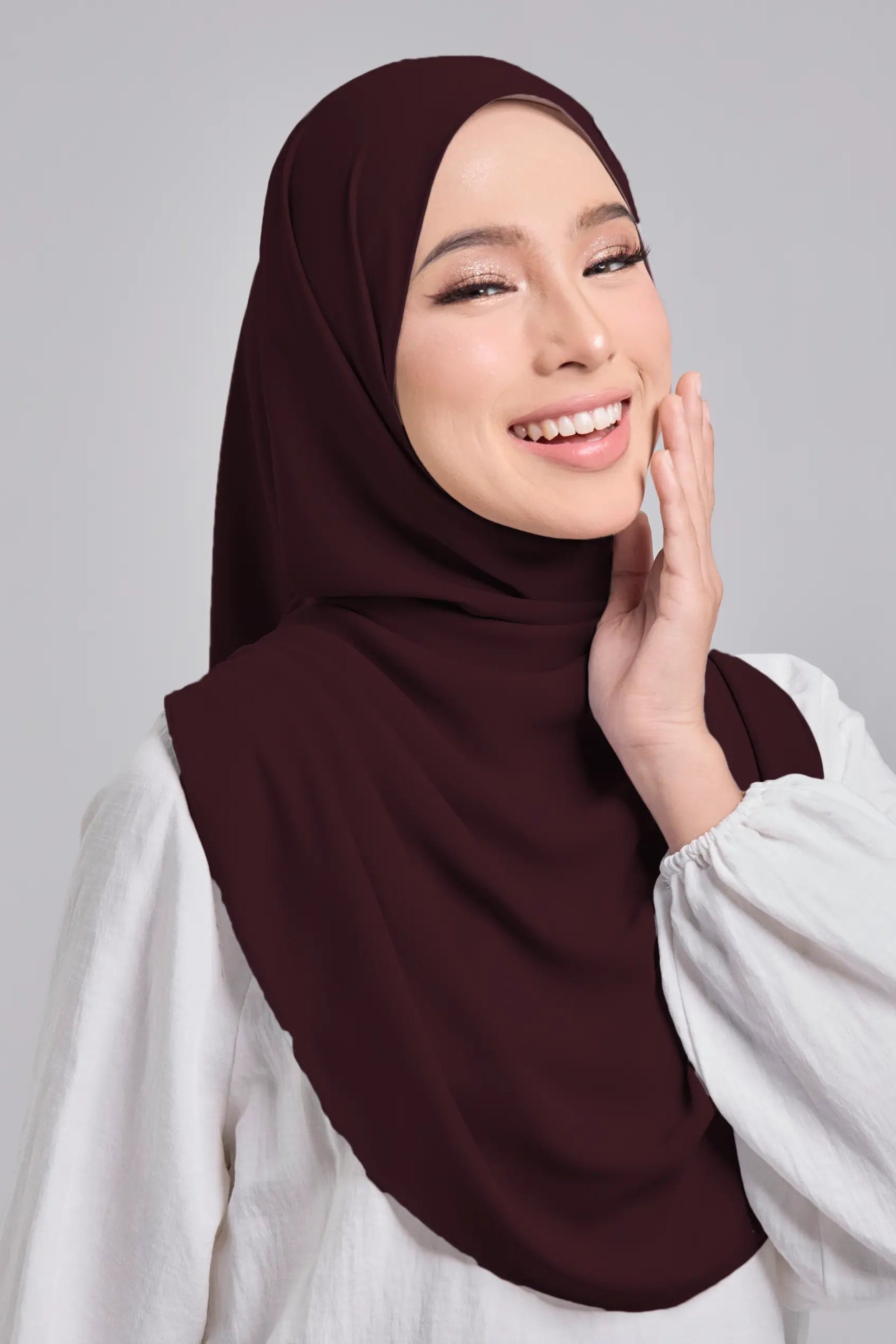 Woman wearing a brown hijab and white shirt against a gray background