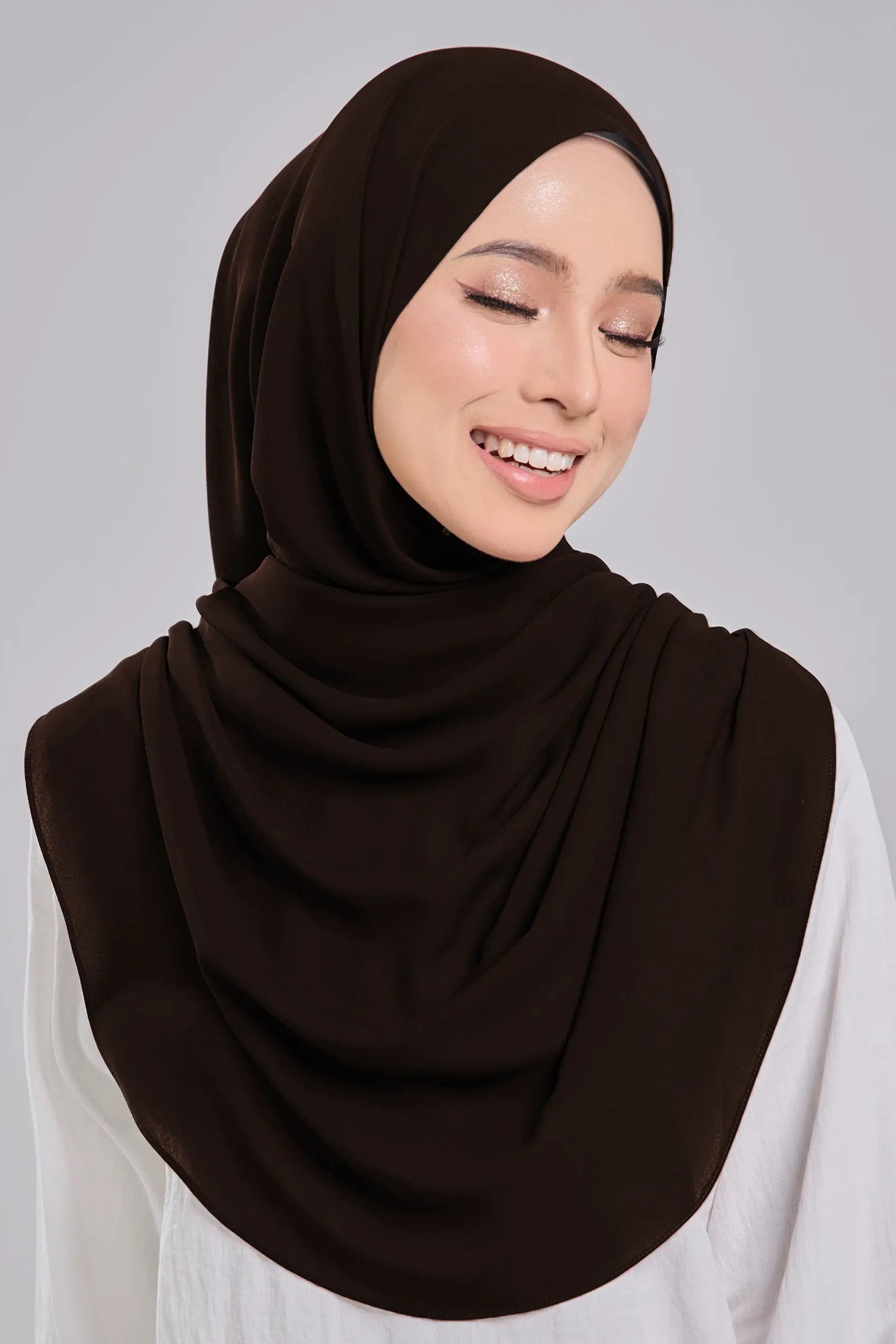 Woman wearing a dark brown hijab and white shirt on a gray background
