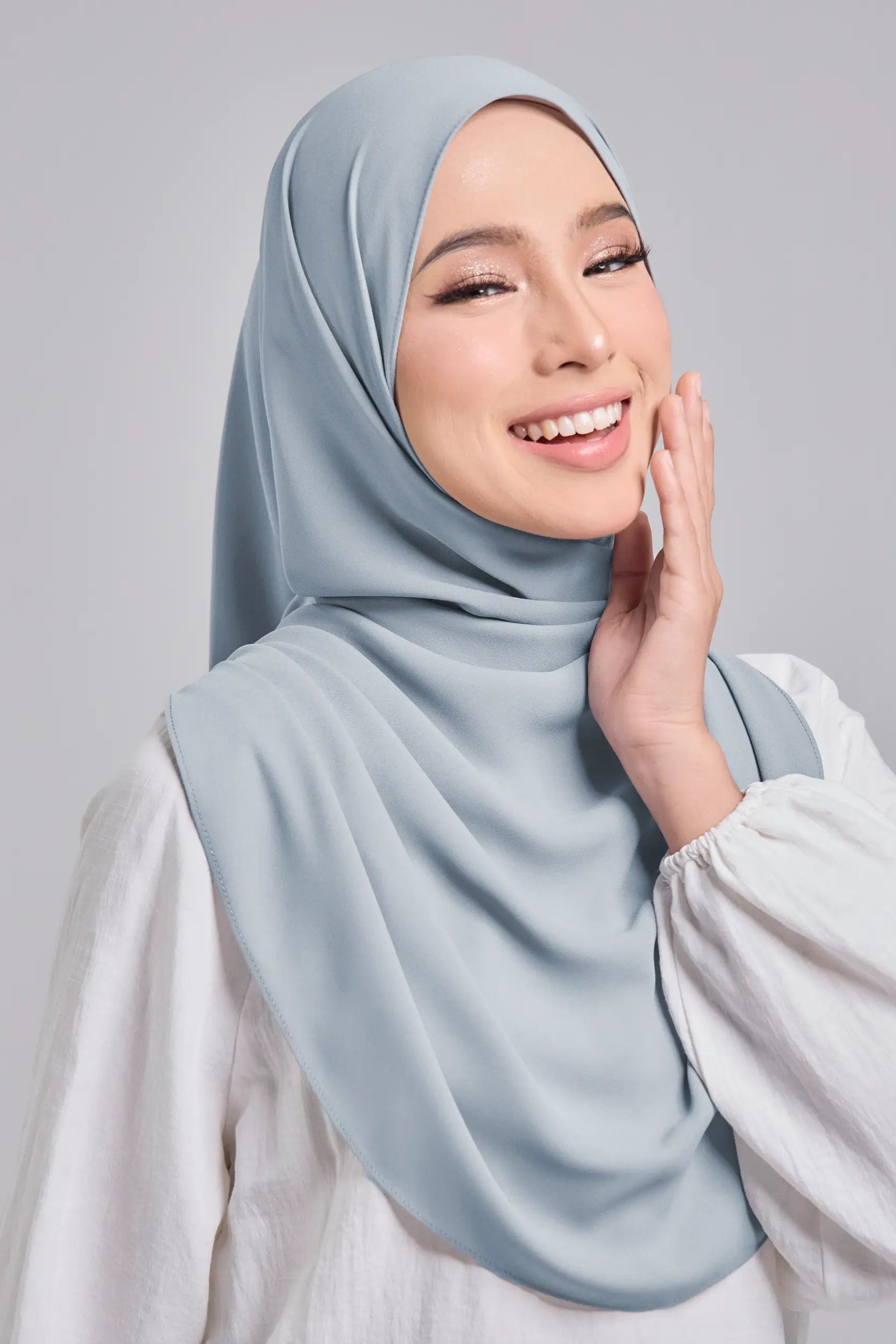 Woman wearing a light blue hijab and white shirt against a gray background