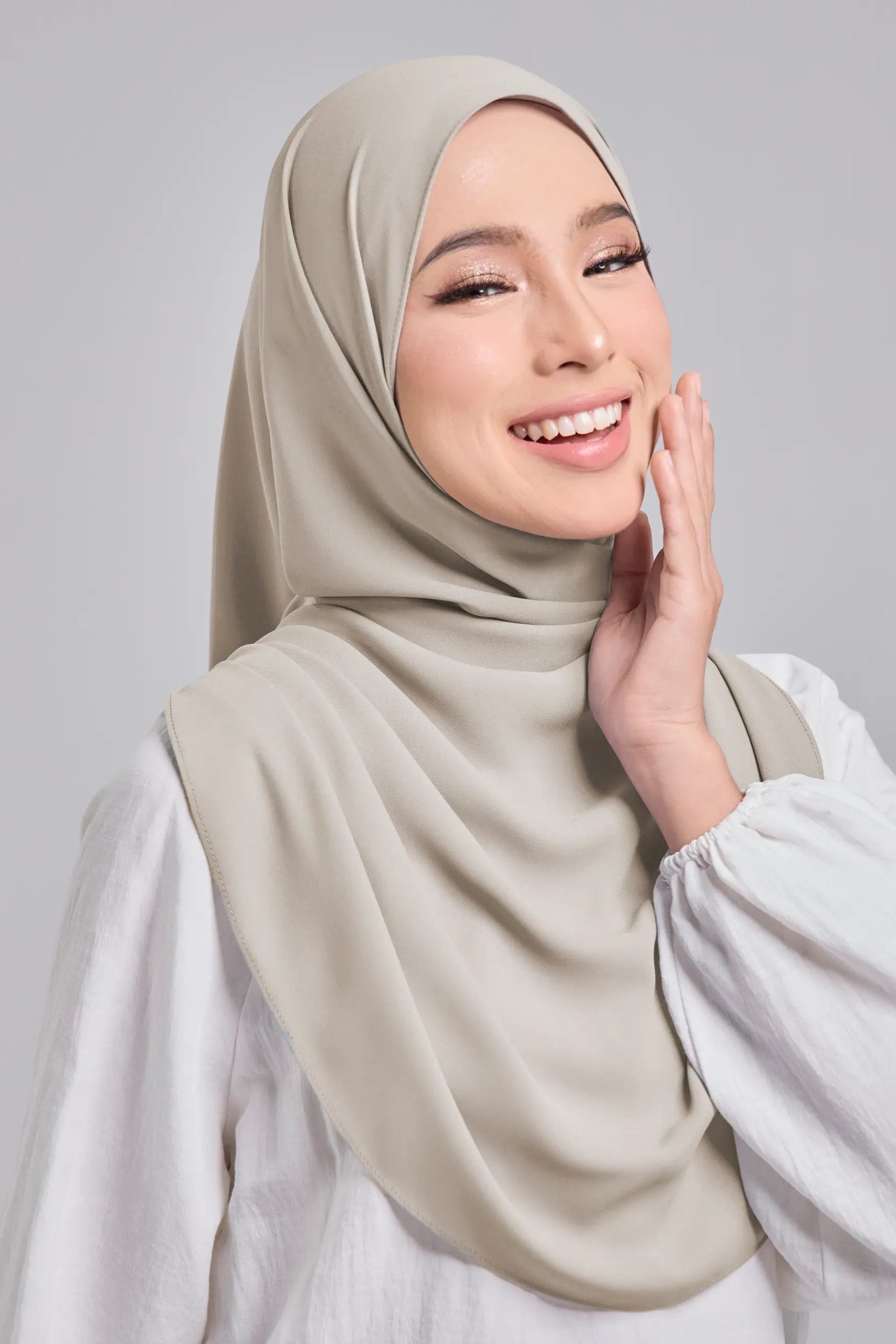 Woman wearing a beige hijab and white top against a gray background