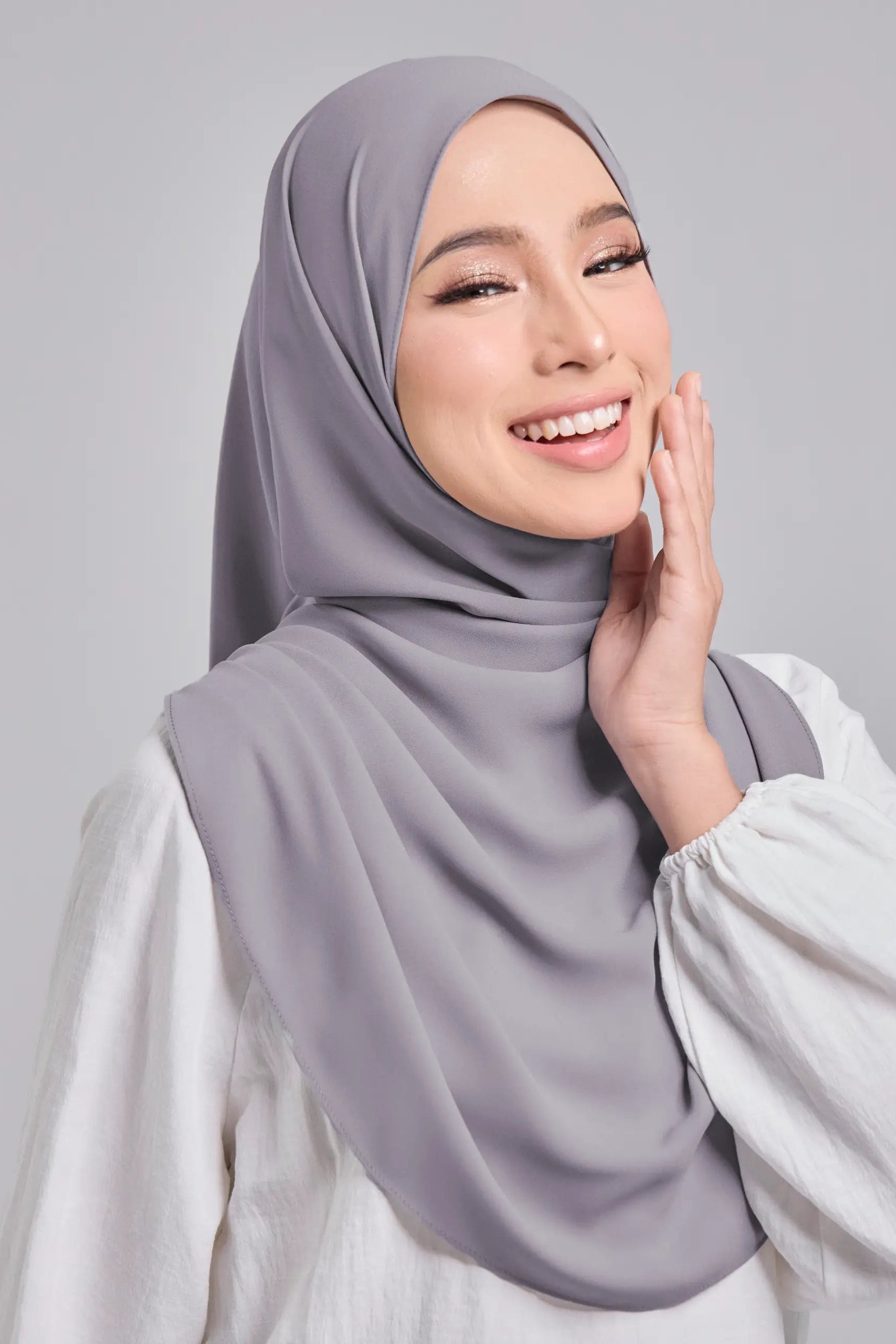 Woman wearing a light gray hijab and white top against a plain background