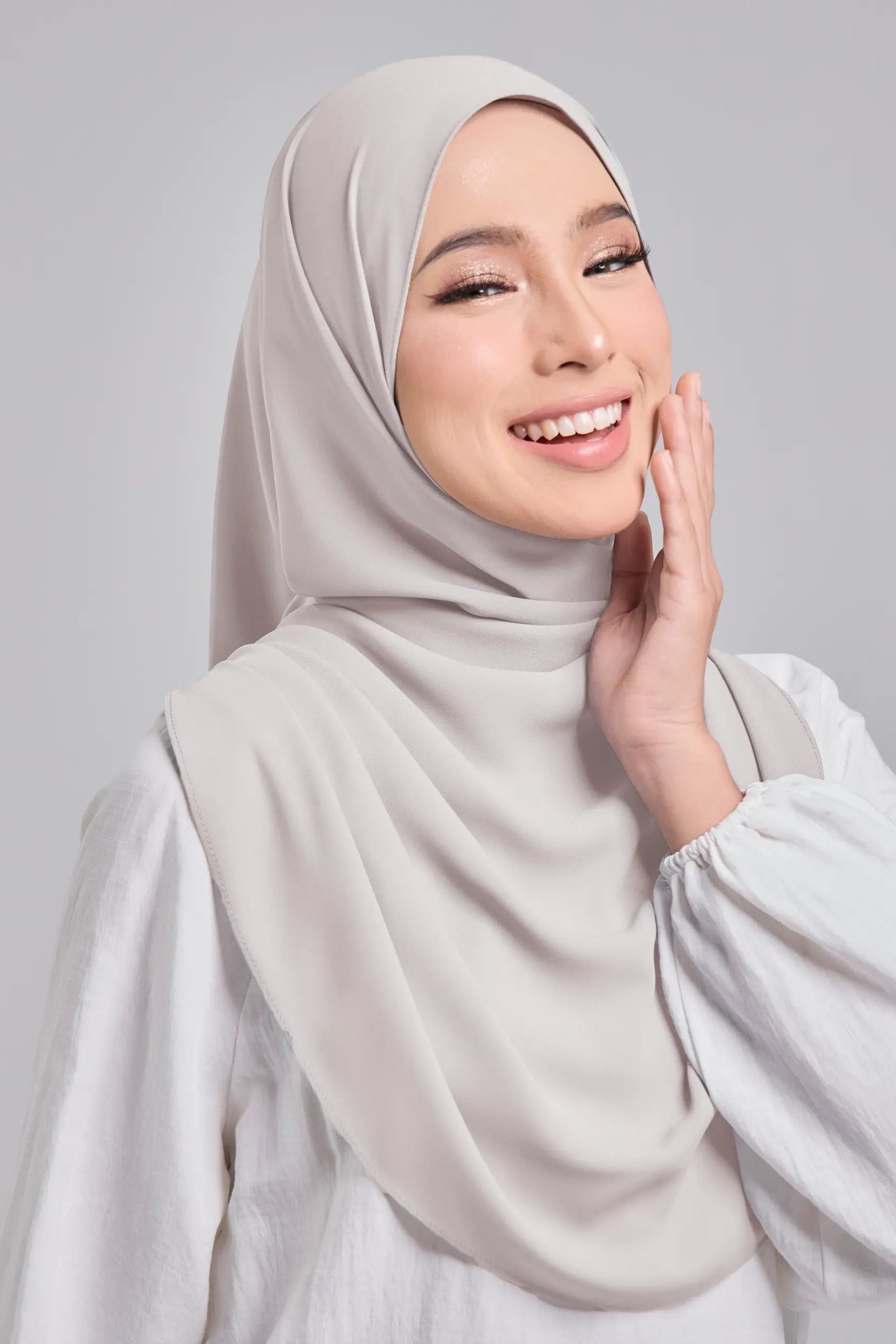Woman wearing a light gray hijab and white top against a plain background