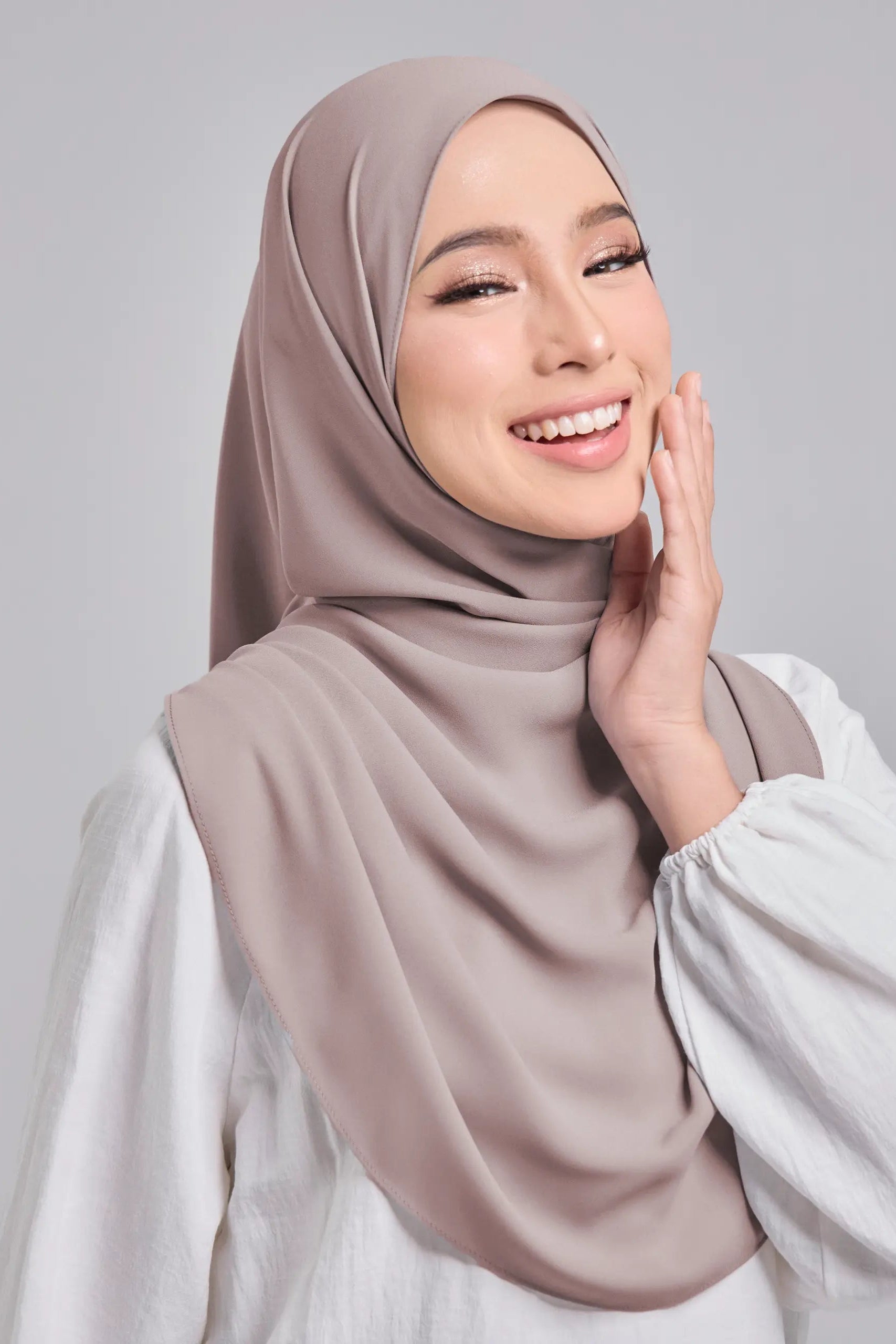 Woman wearing a beige hijab and white top against a gray background