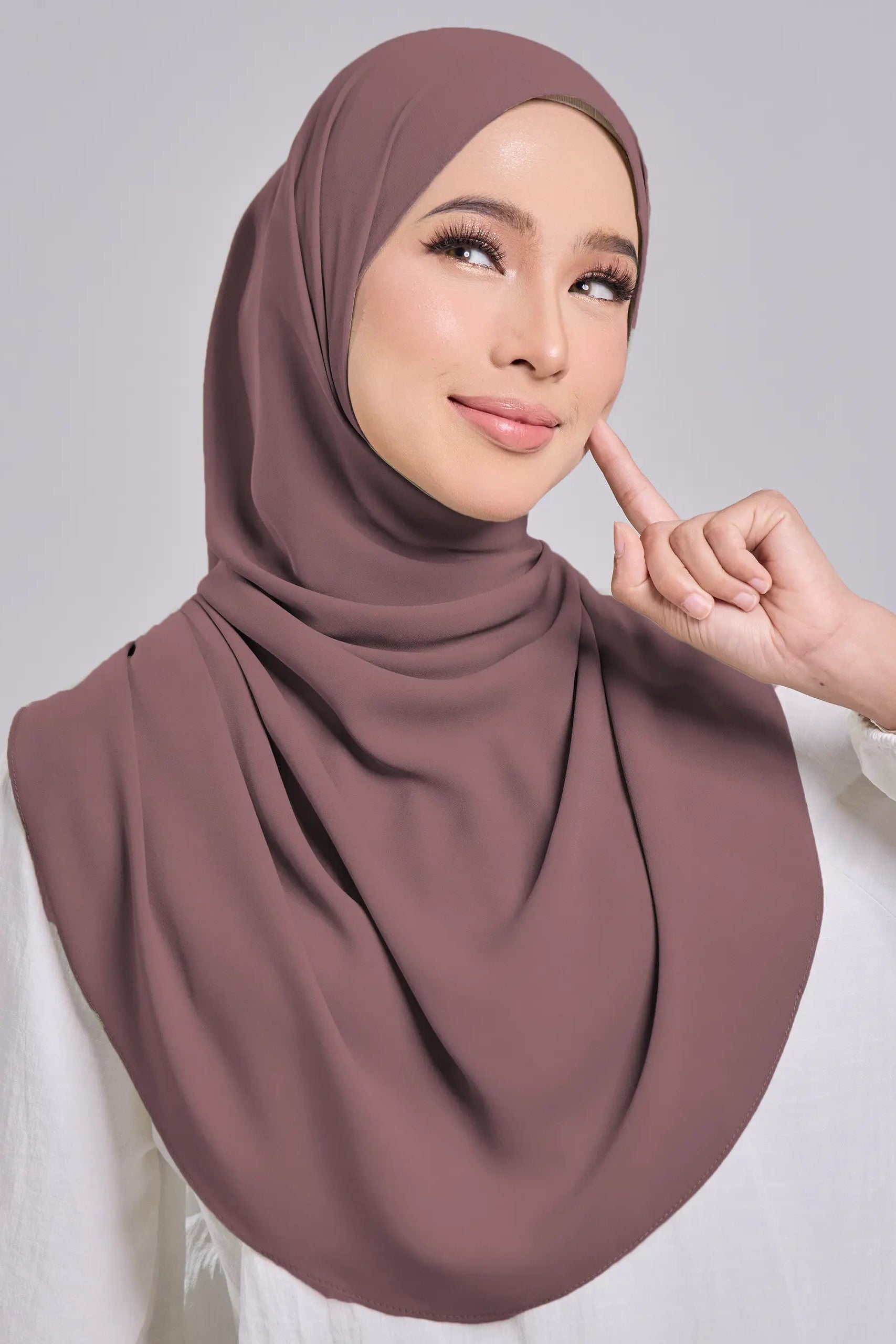 Woman wearing a brown hijab and white top against a gray background
