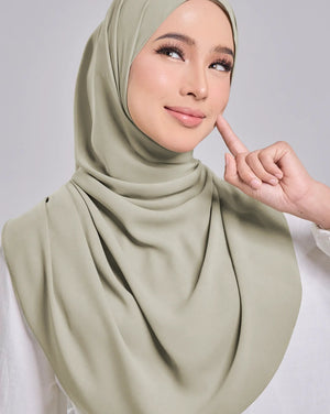 A woman wearing a draped sage green hijab and a white long-sleeve top, posing against a light gray studio background.