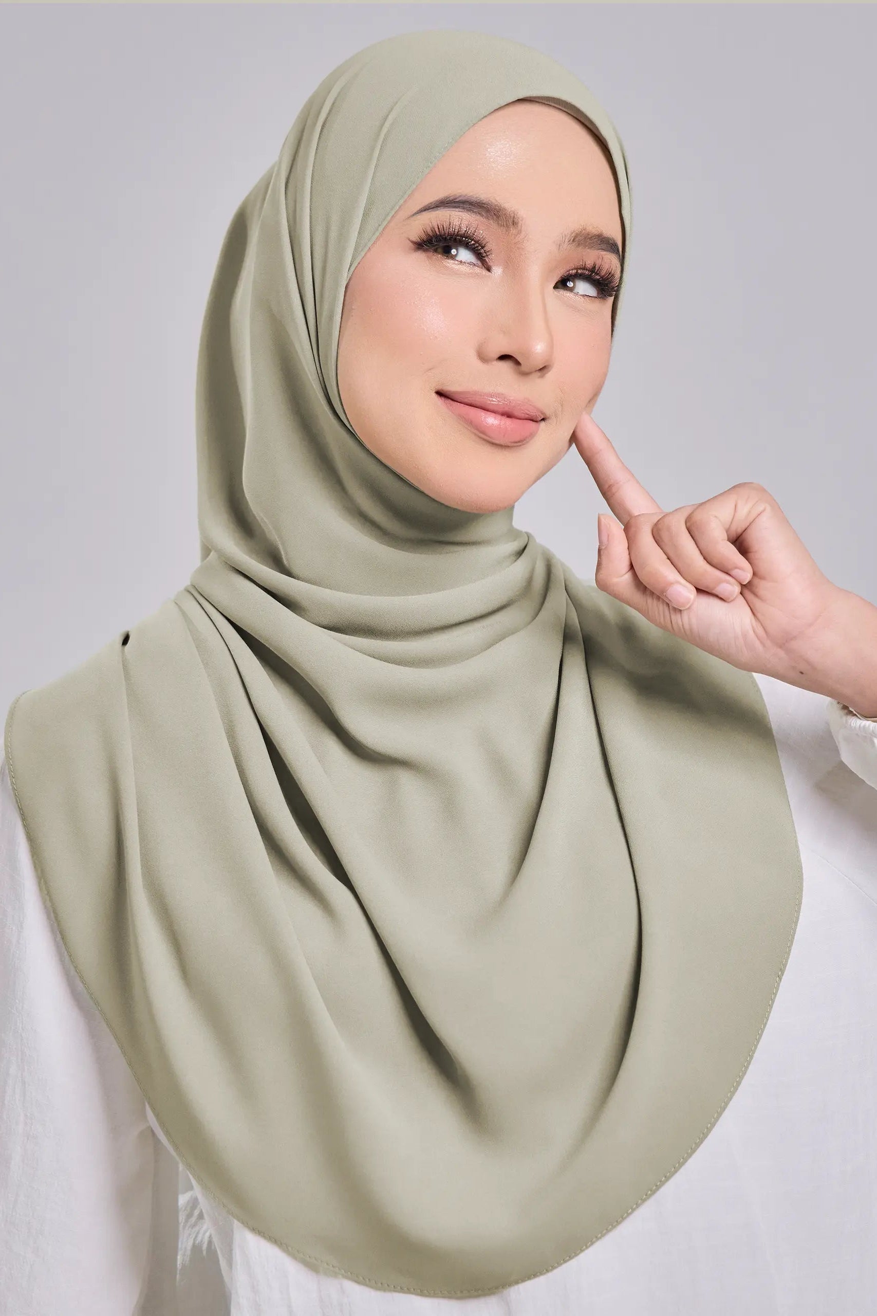 A woman wearing a draped sage green hijab and a white long-sleeve top, posing against a light gray studio background.