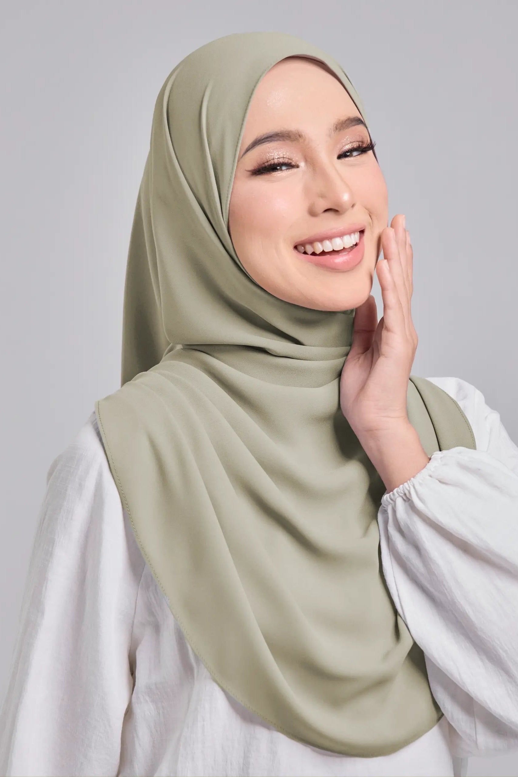 A woman wearing a soft sage green hijab draped elegantly over her shoulders, paired with a white long-sleeve blouse, set against a solid light gray studio background.