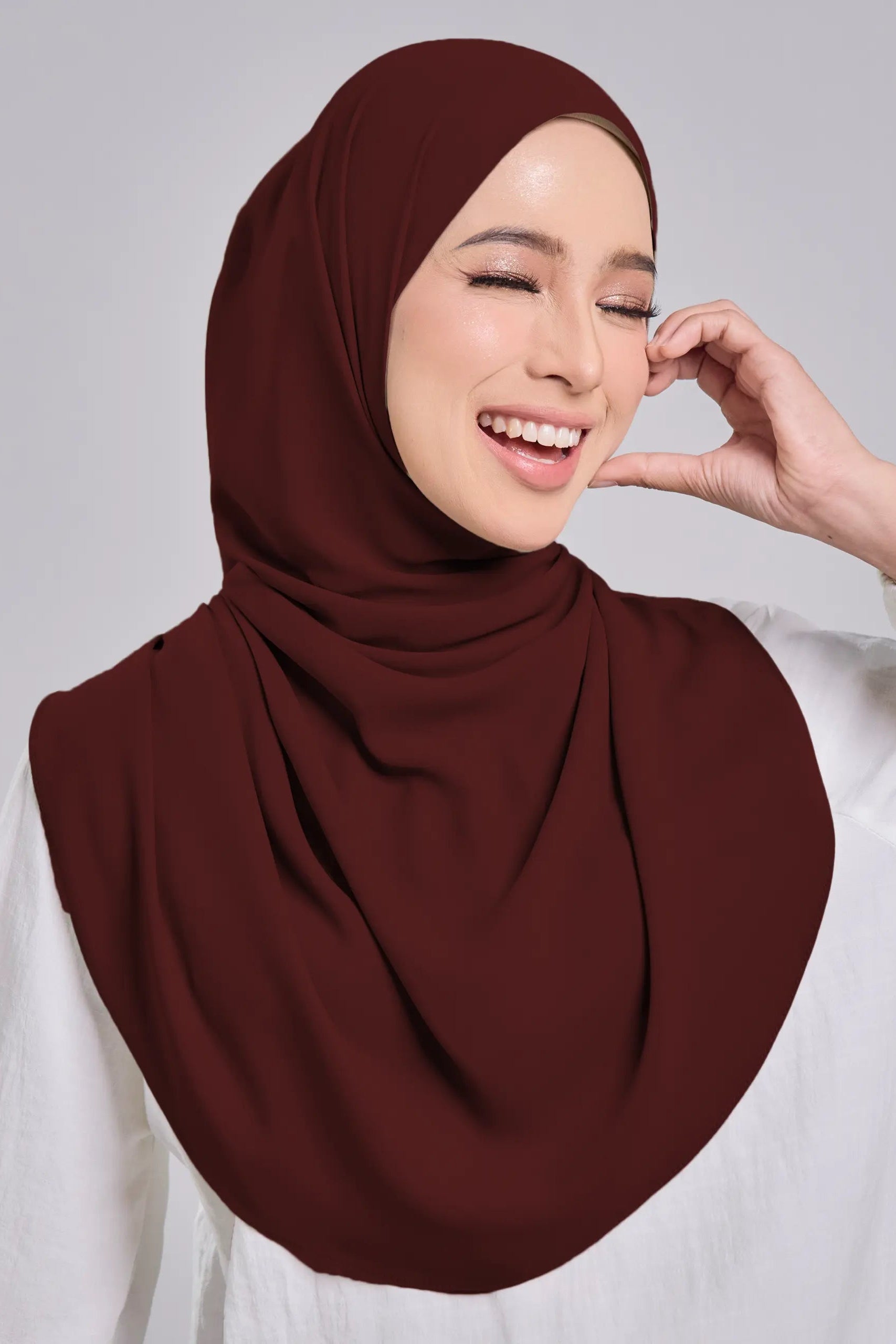 Woman wearing a maroon hijab and white shirt on a gray background