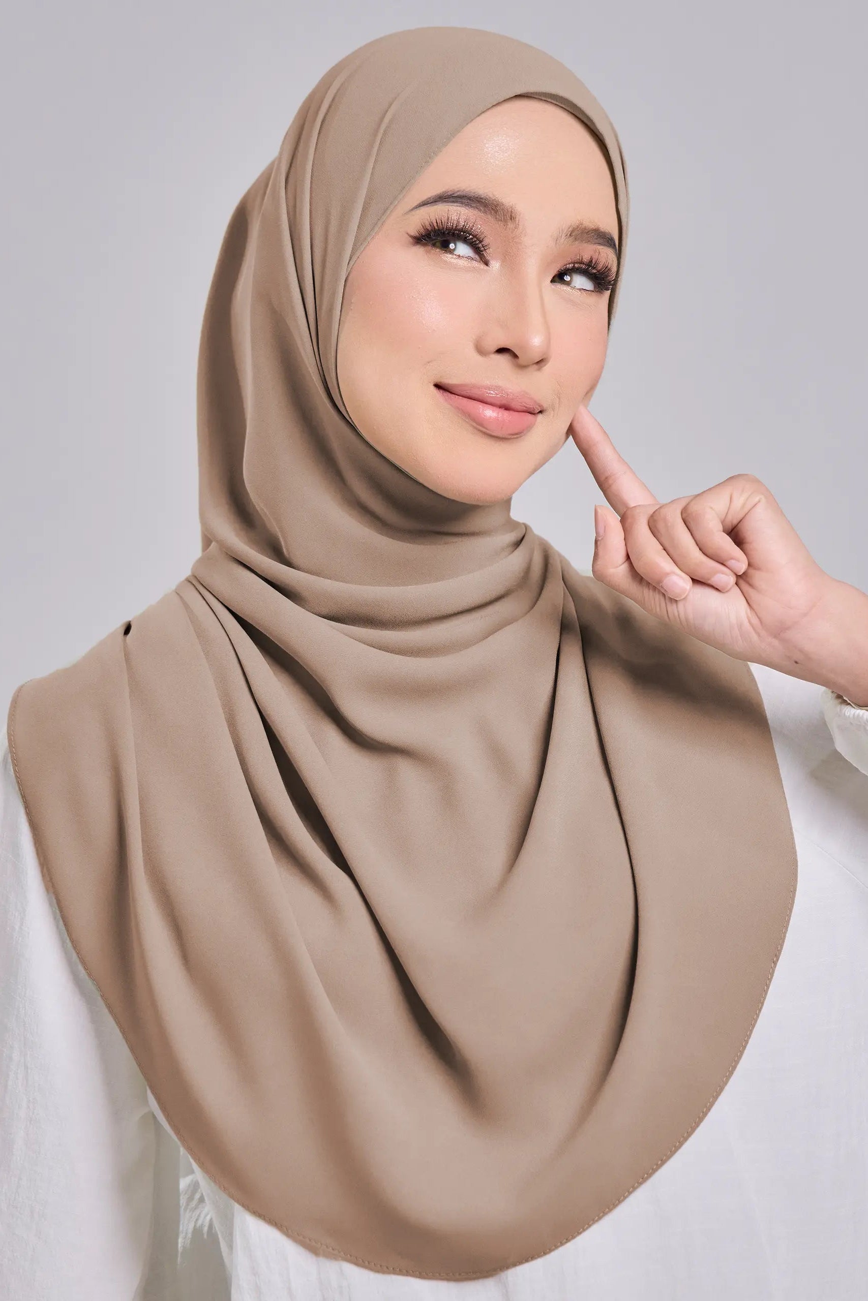 Woman wearing a beige hijab and white top on a gray background