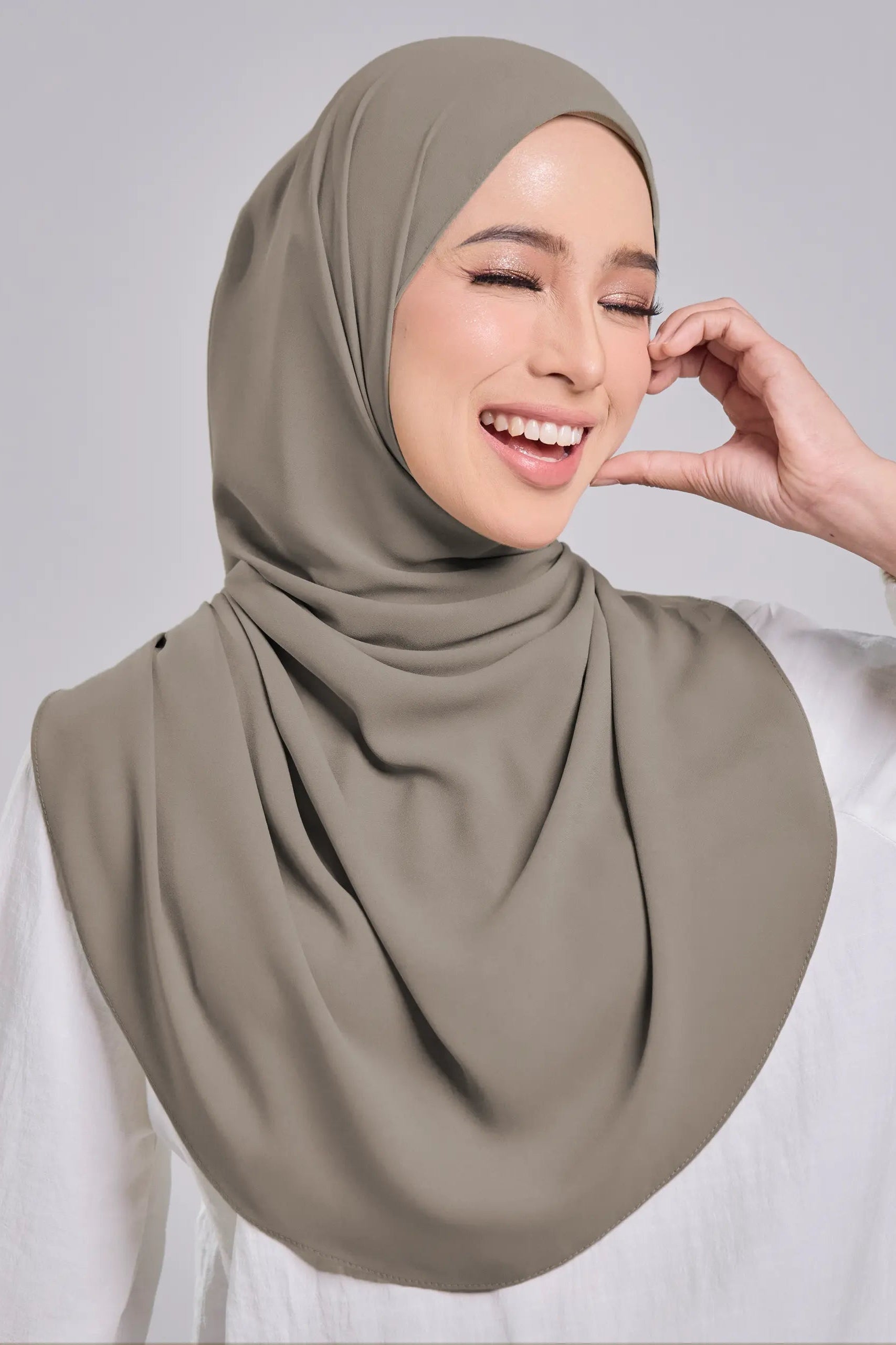 A woman wearing a draped taupe-gray hijab and a white long-sleeve top, smiling against a solid light gray background.