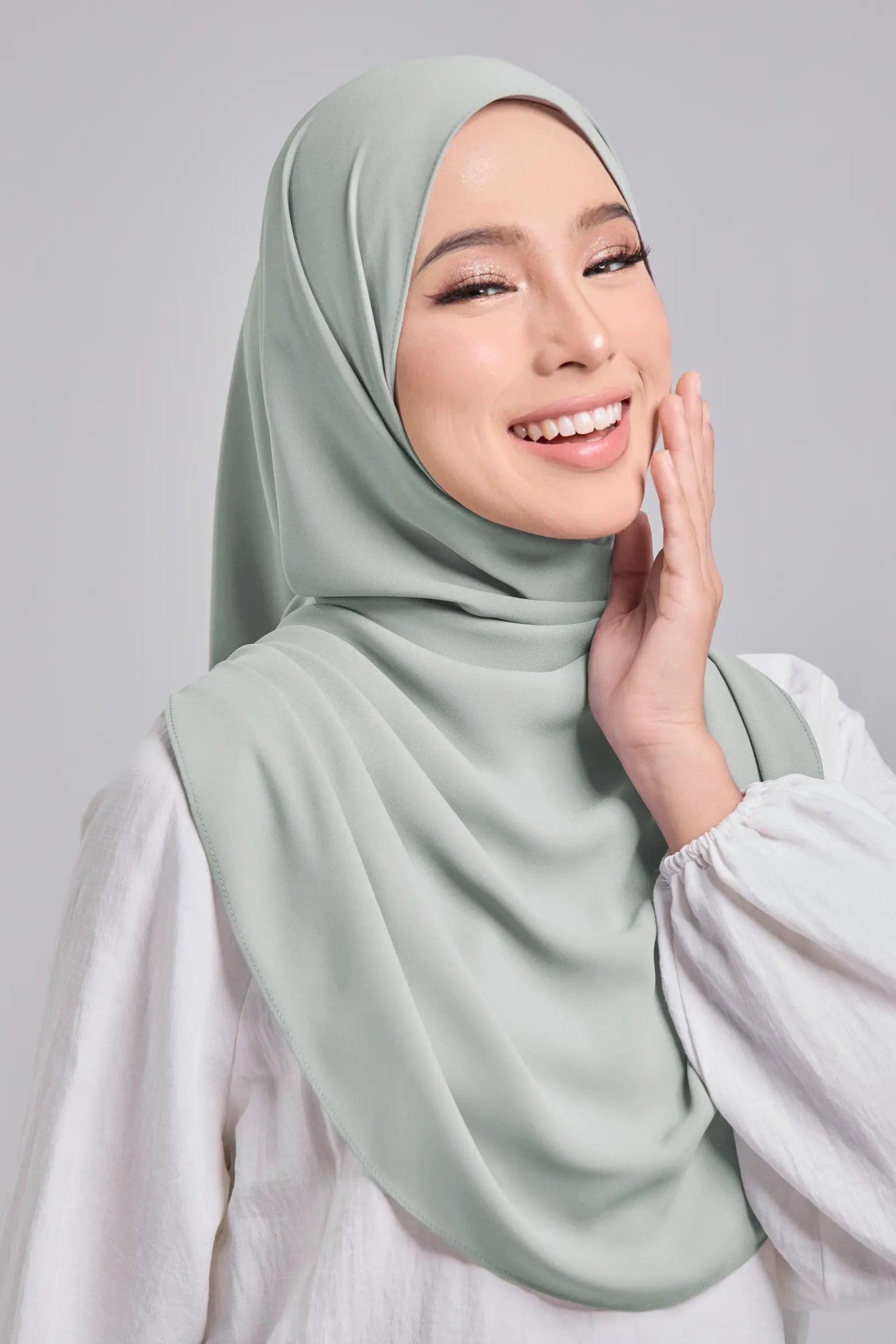 Woman wearing a light green hijab and white top against a gray background