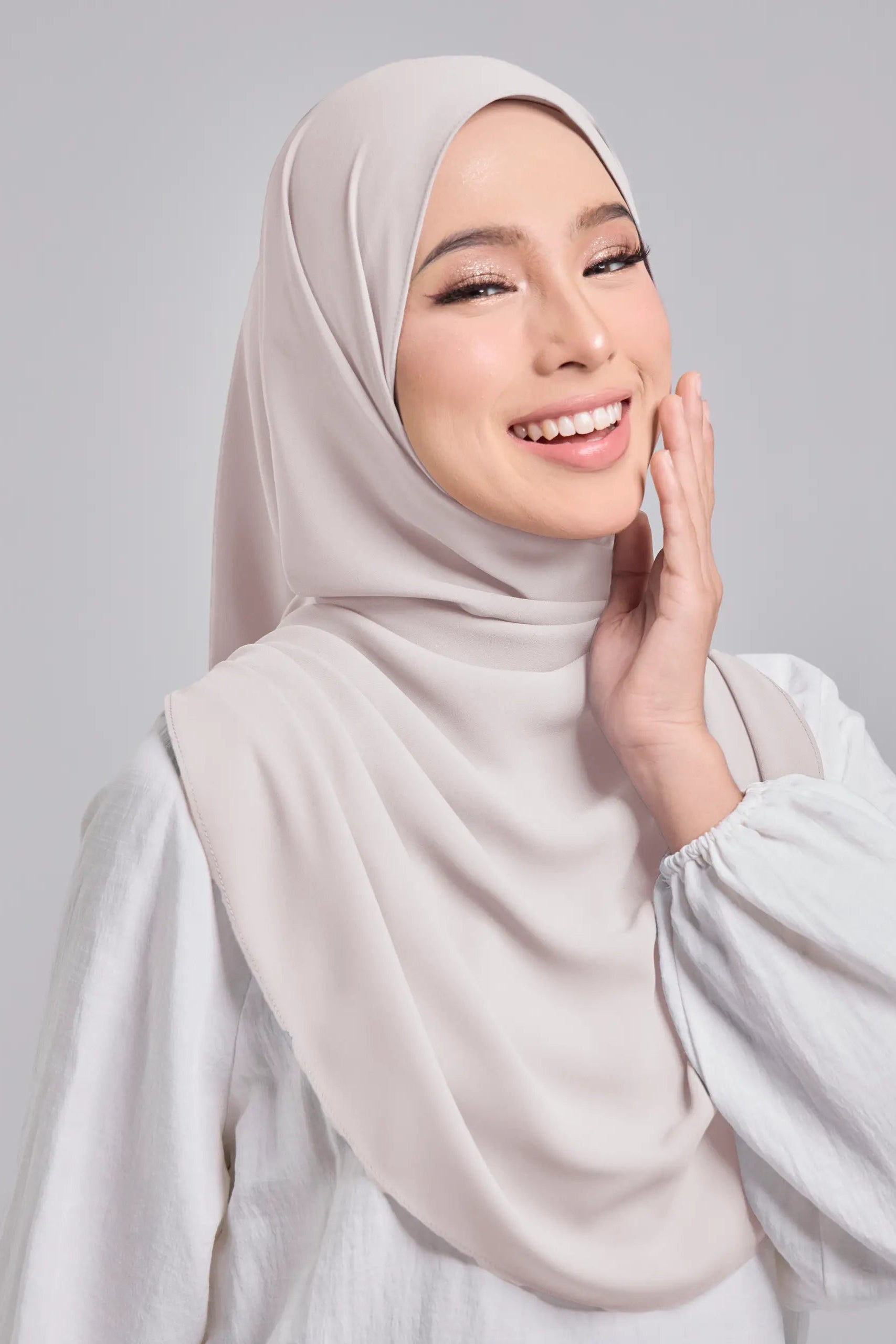 Woman wearing a beige hijab and white top against a light gray background