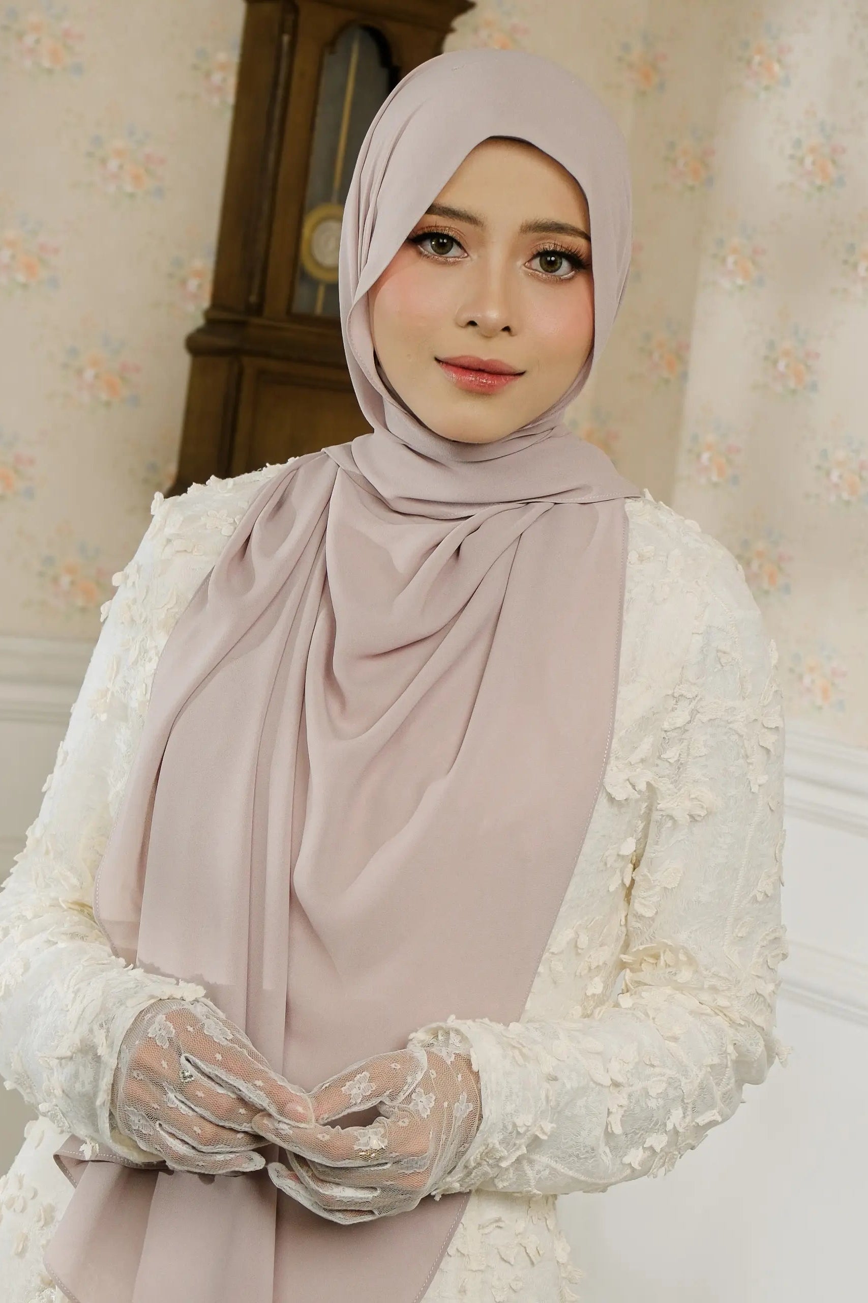Woman wearing a light pink hijab and white lace garment in a decorated room.