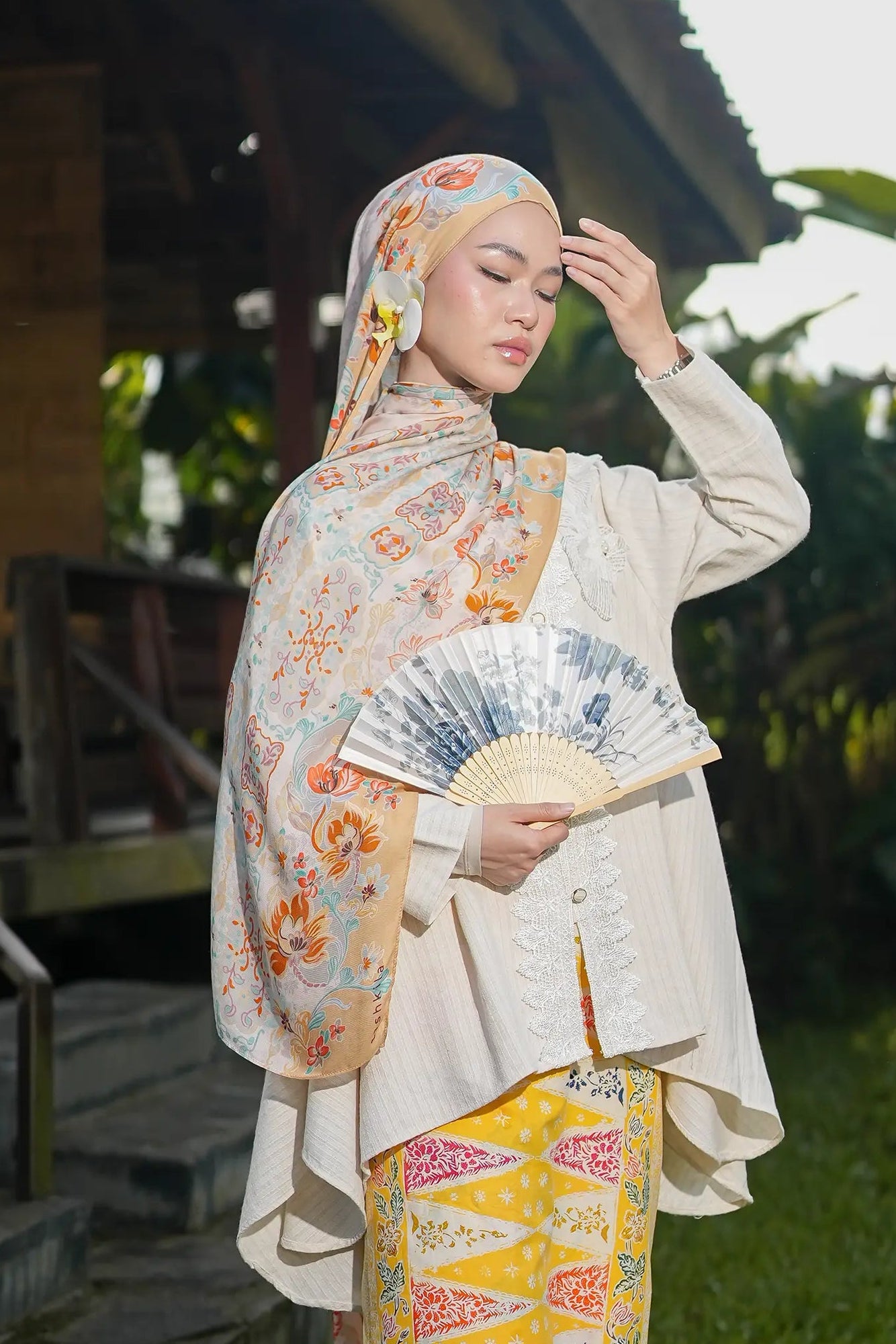 Woman in traditional outfit holding a fan outdoors with greenery around