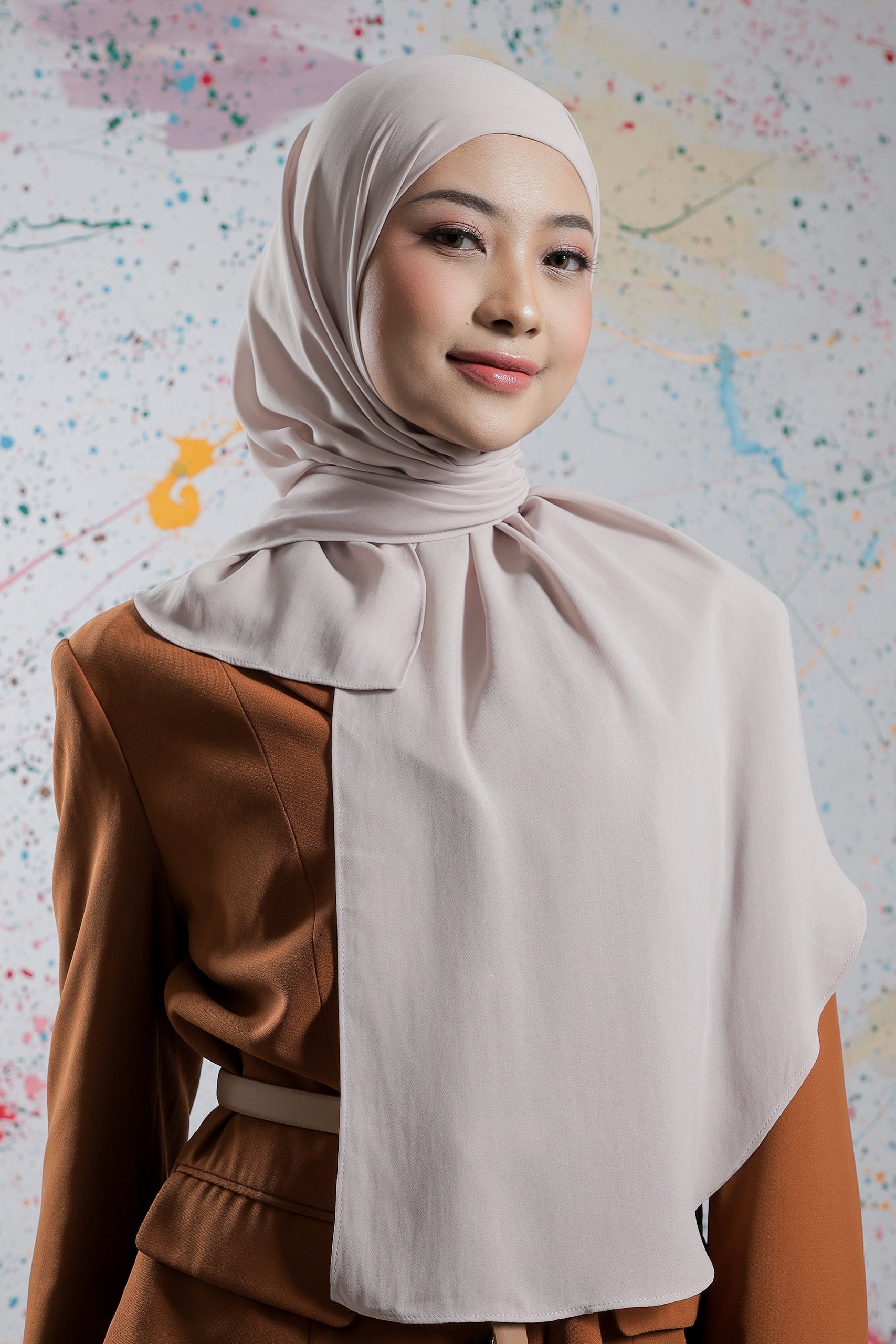 Woman wearing a beige hijab and brown outfit against a colorful abstract background