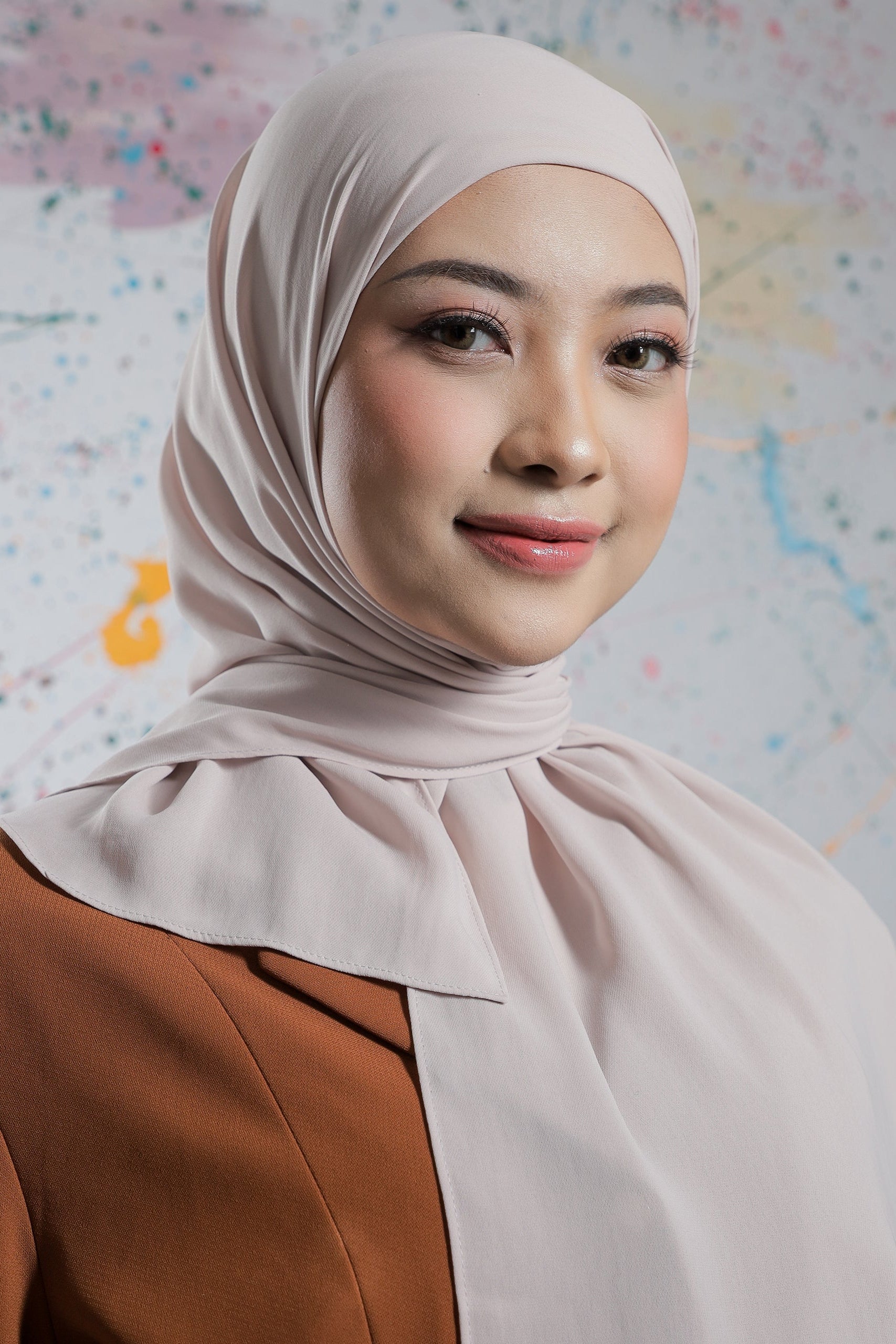 Woman wearing a beige hijab and brown top against a colorful abstract background