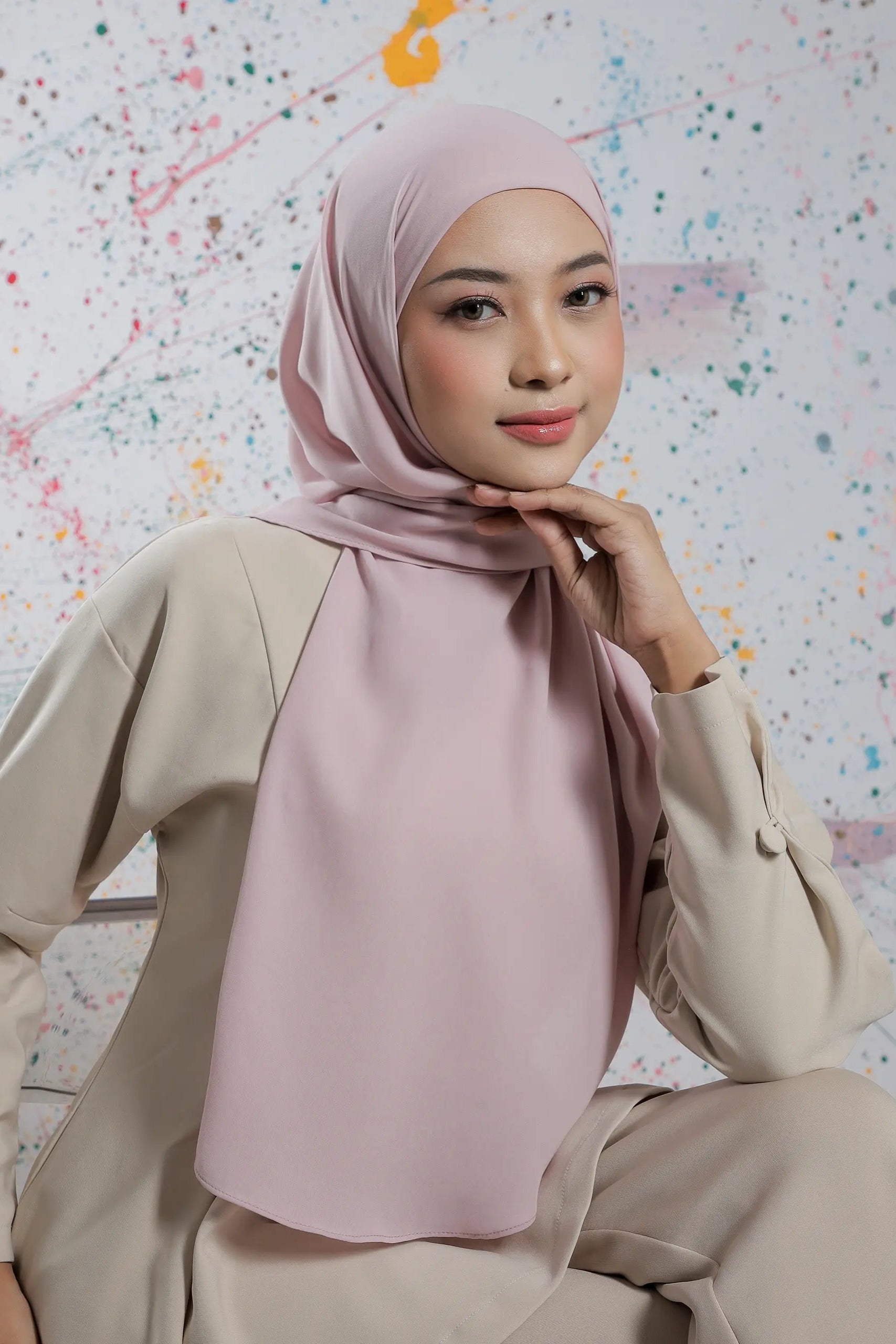 Woman wearing a light pink hijab and beige outfit against a colorful abstract background