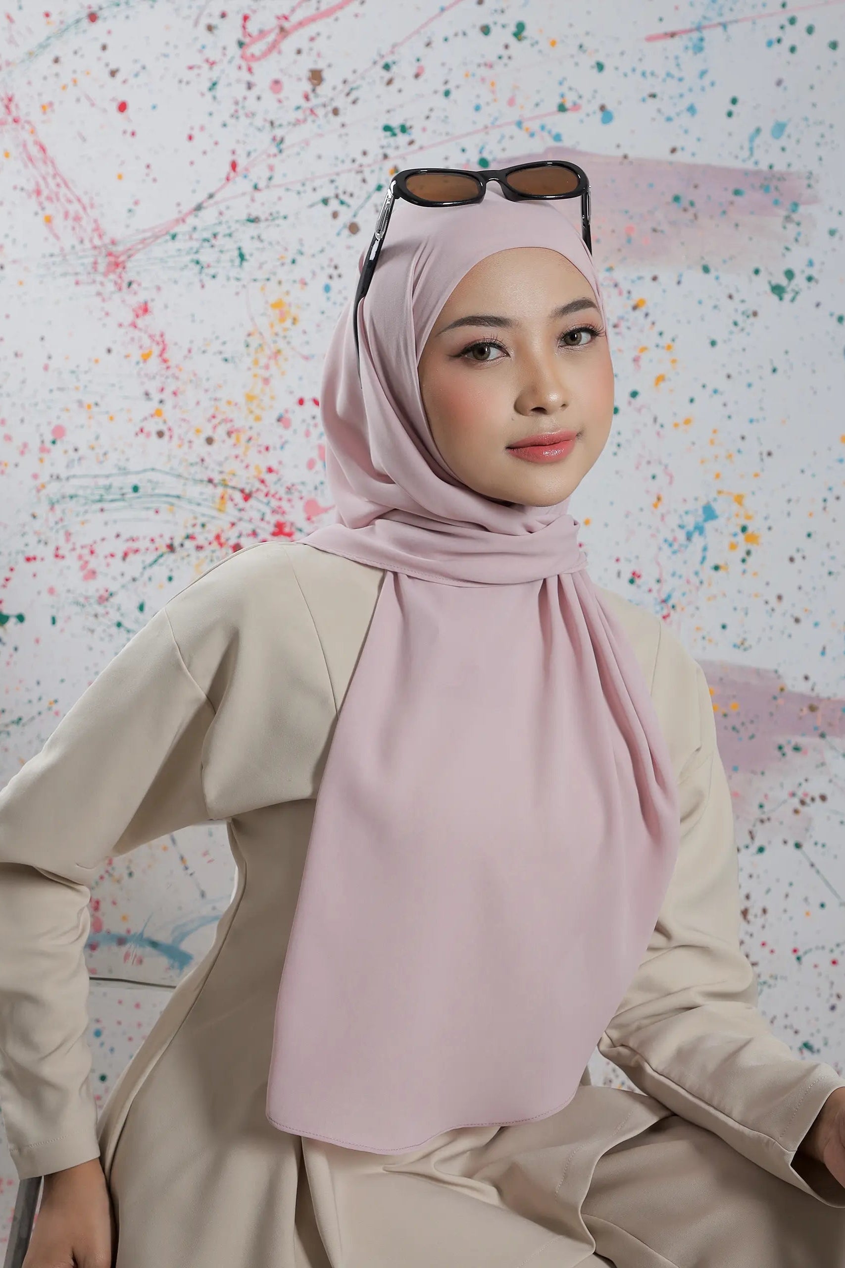 Woman wearing a pink hijab and beige outfit against a colorful abstract background