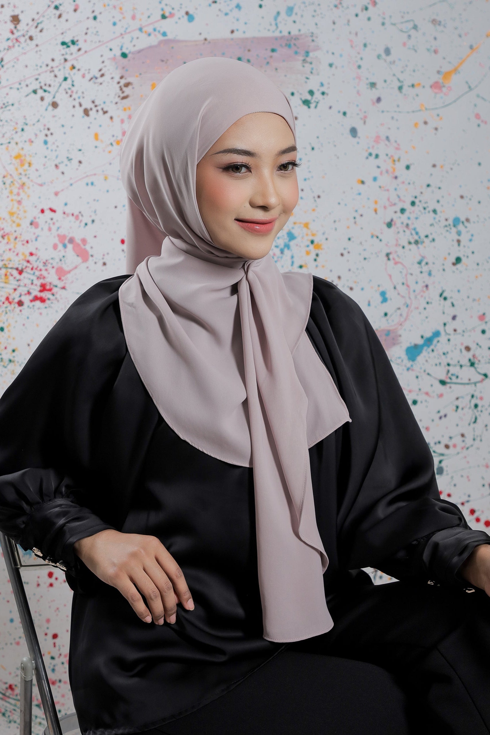 Woman wearing a light pink hijab and black outfit against a colorful abstract background