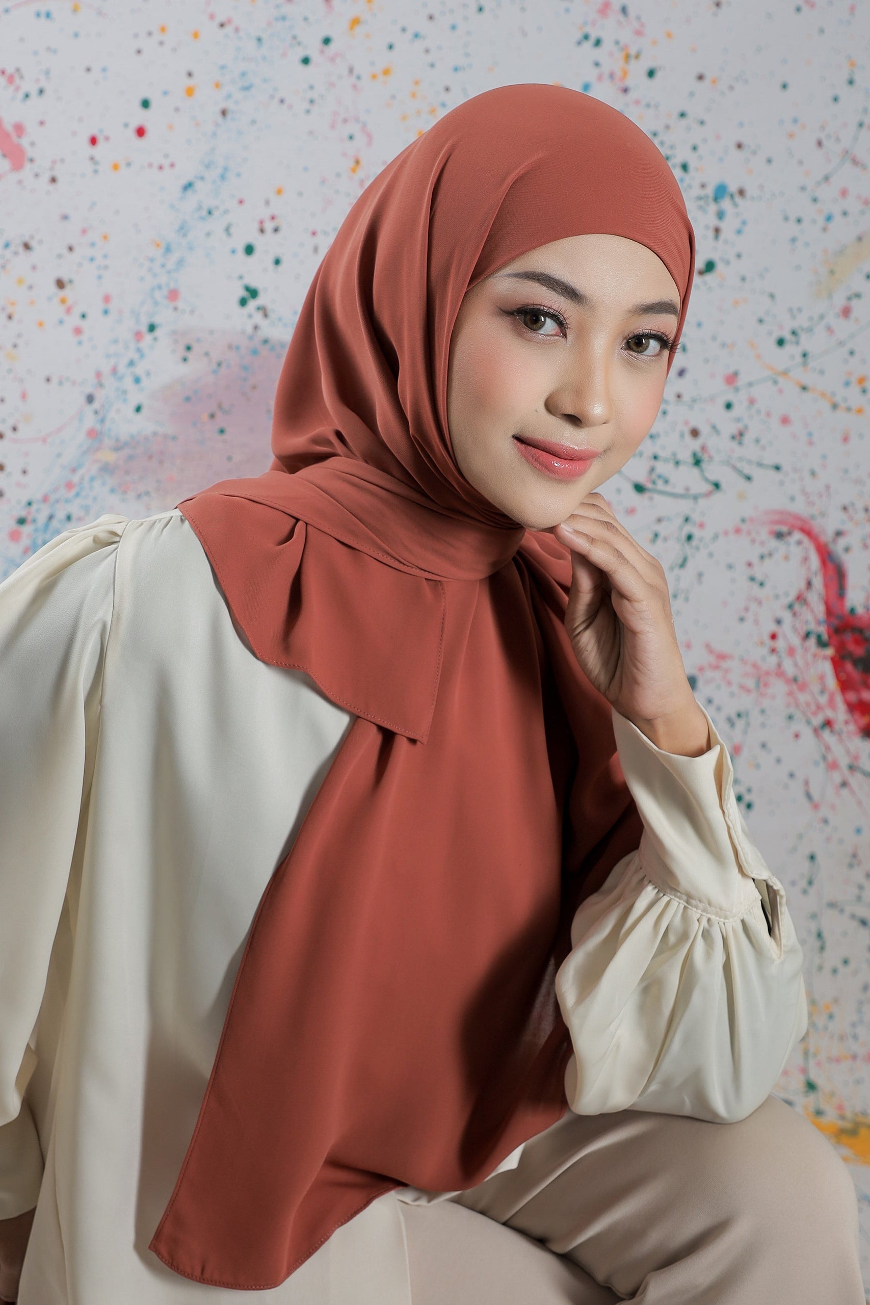 Woman wearing a rust-colored hijab and beige outfit against a colorful abstract background