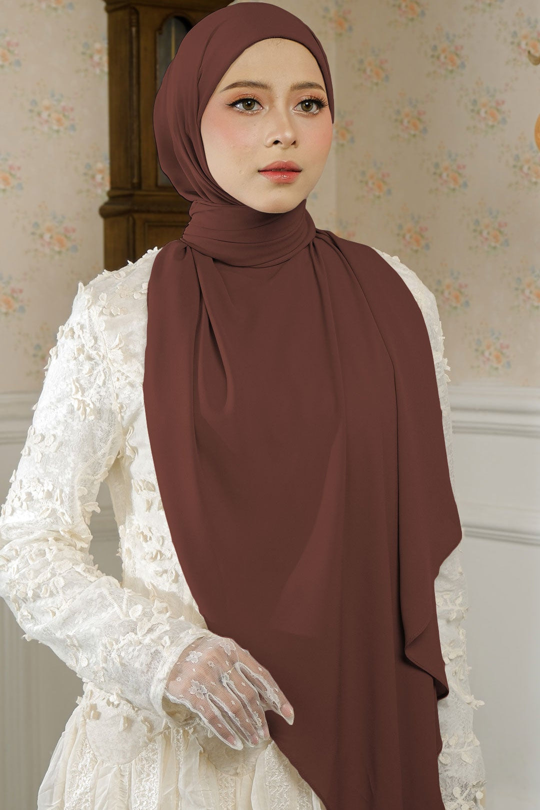 Woman wearing a brown hijab and white lace dress in a decorated room.