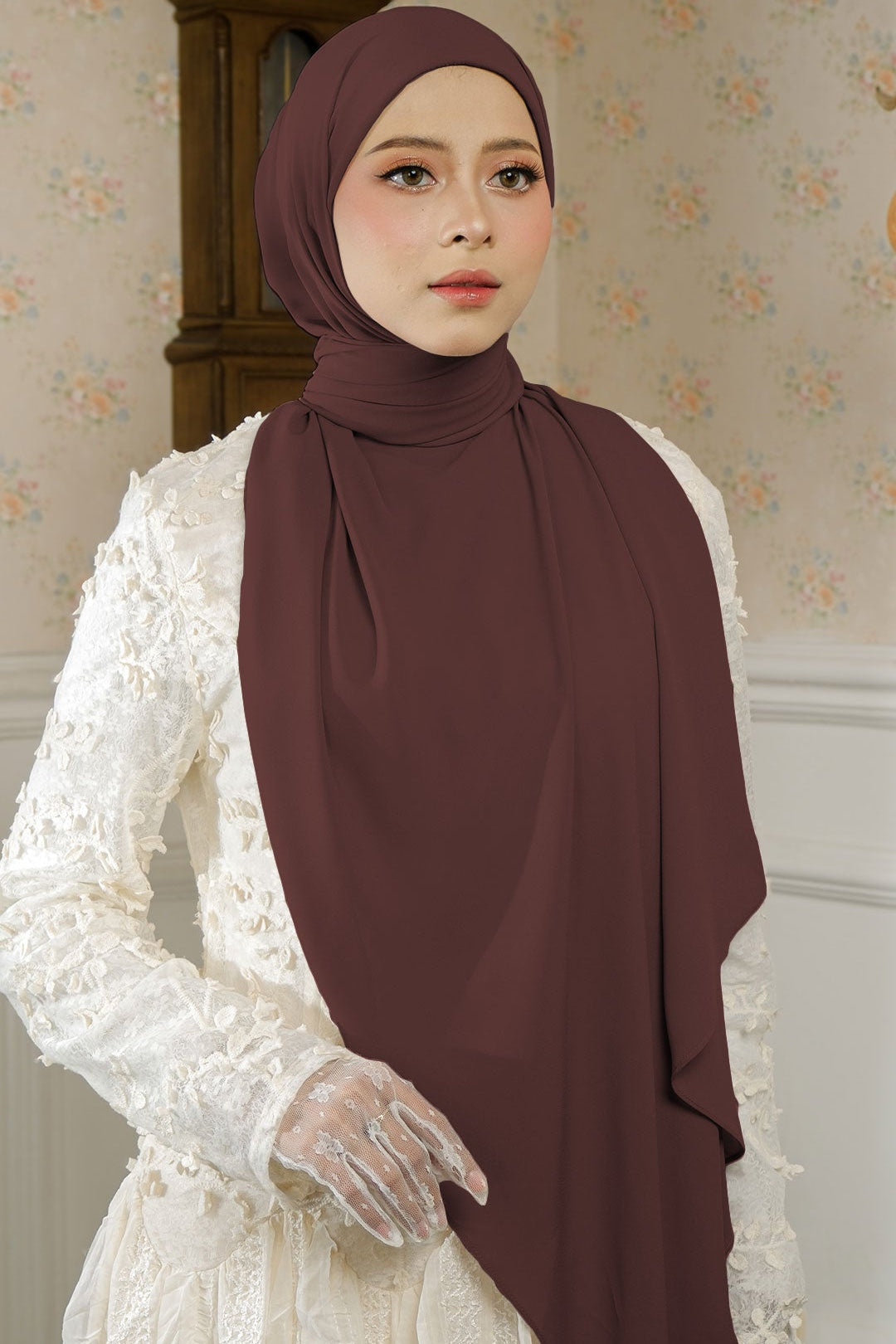 Woman wearing a brown hijab and white lace dress in a decorated room.