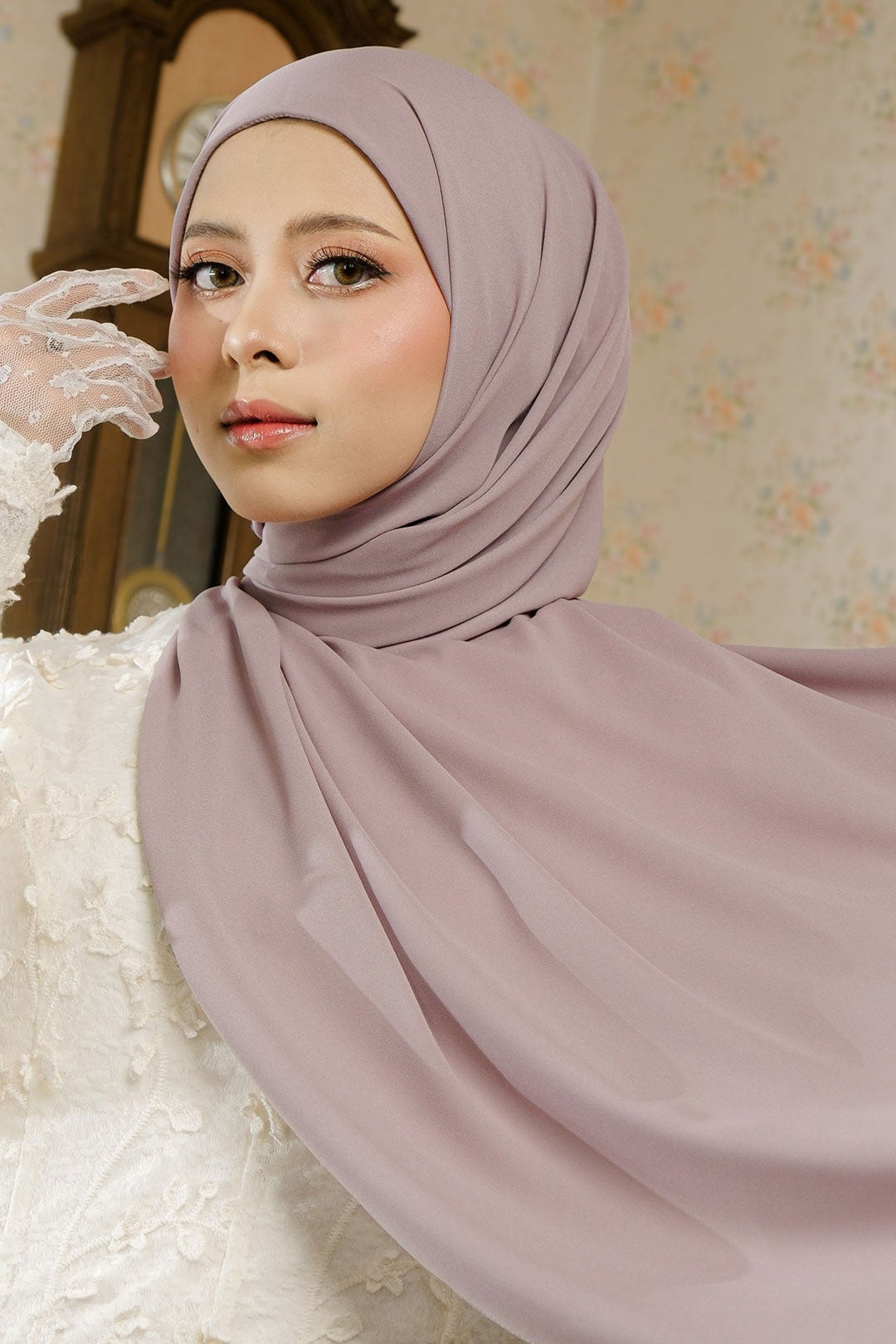 Woman wearing a light purple hijab in a room with floral wallpaper.