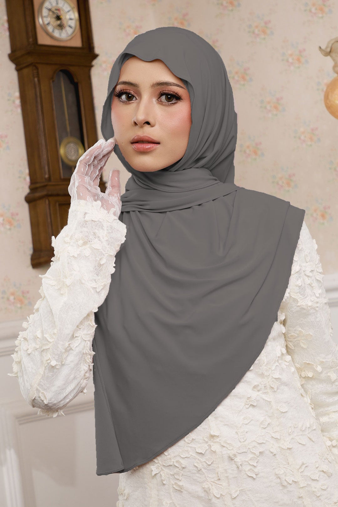 Woman wearing a gray hijab and white gloves in a decorative setting with a clock in the background.