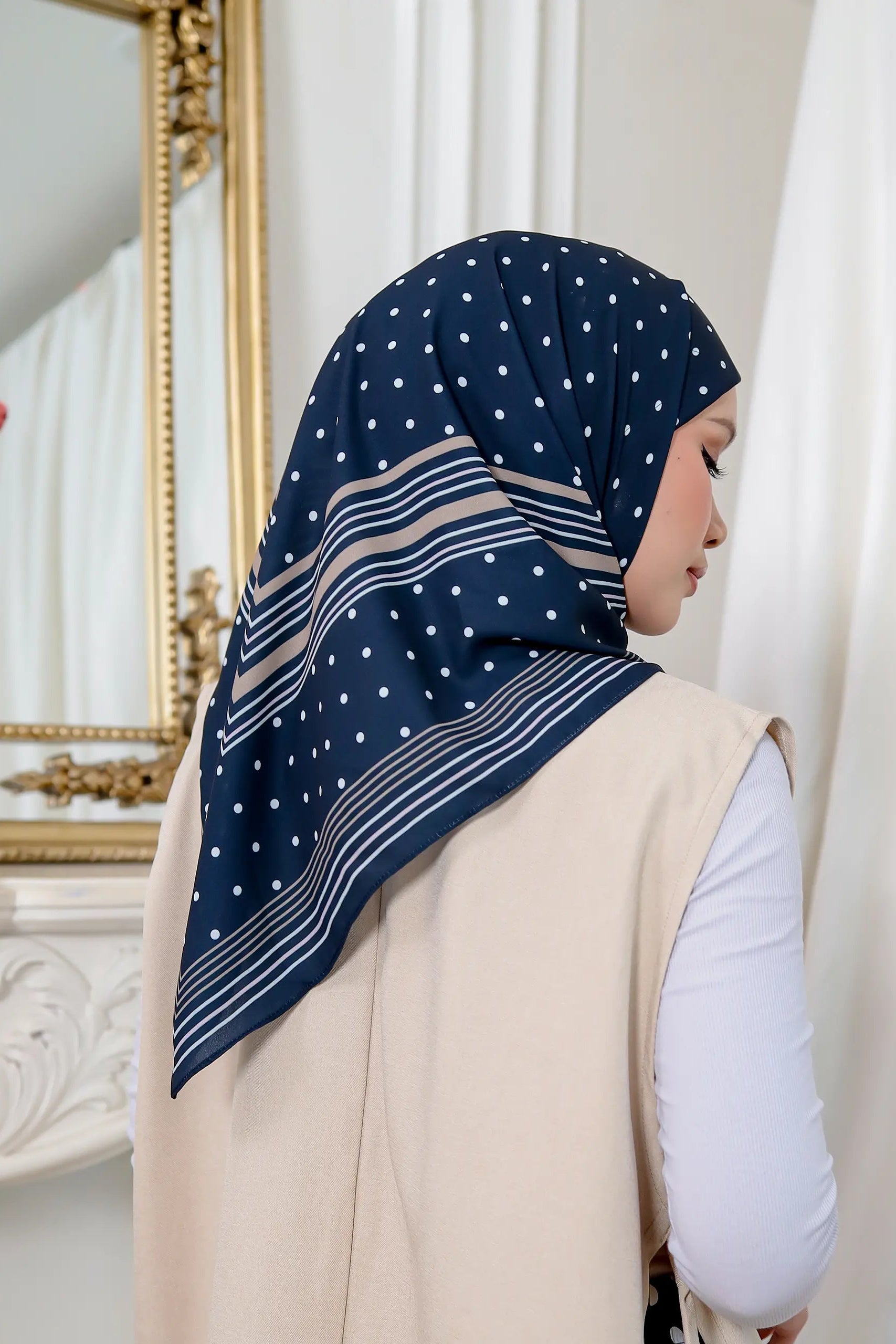 Woman wearing a navy hijab with white polka dots and stripes, standing in front of a mirror.