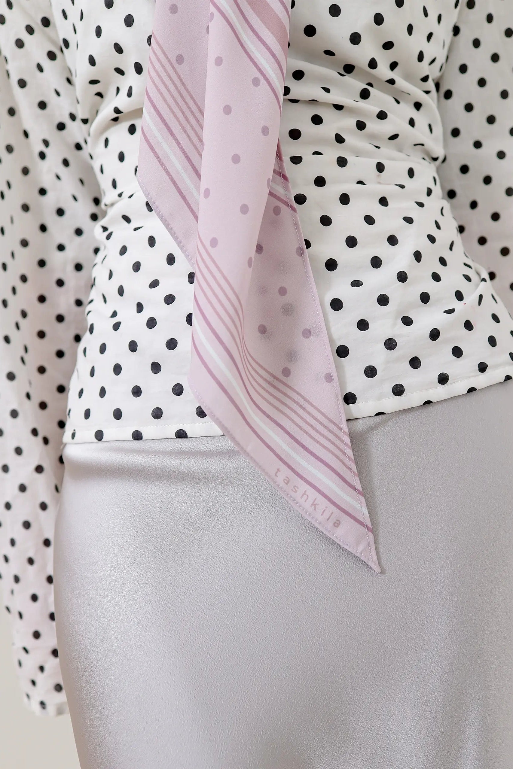 Woman wearing a polka dot blouse with a pink scarf and light gray skirt.