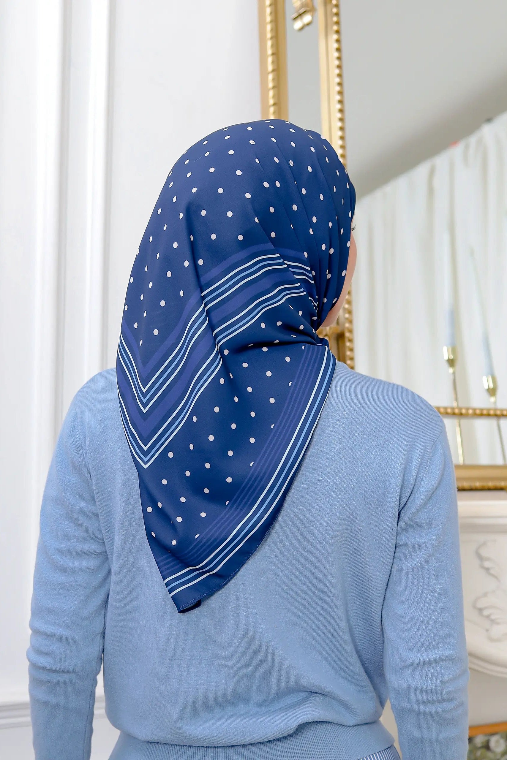 Person wearing a blue headscarf with white patterns in a room with white curtains and a decorative mirror.
