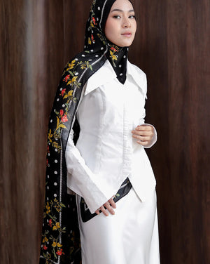 Woman wearing a white outfit with a black hijab featuring floral embroidery against a brown background