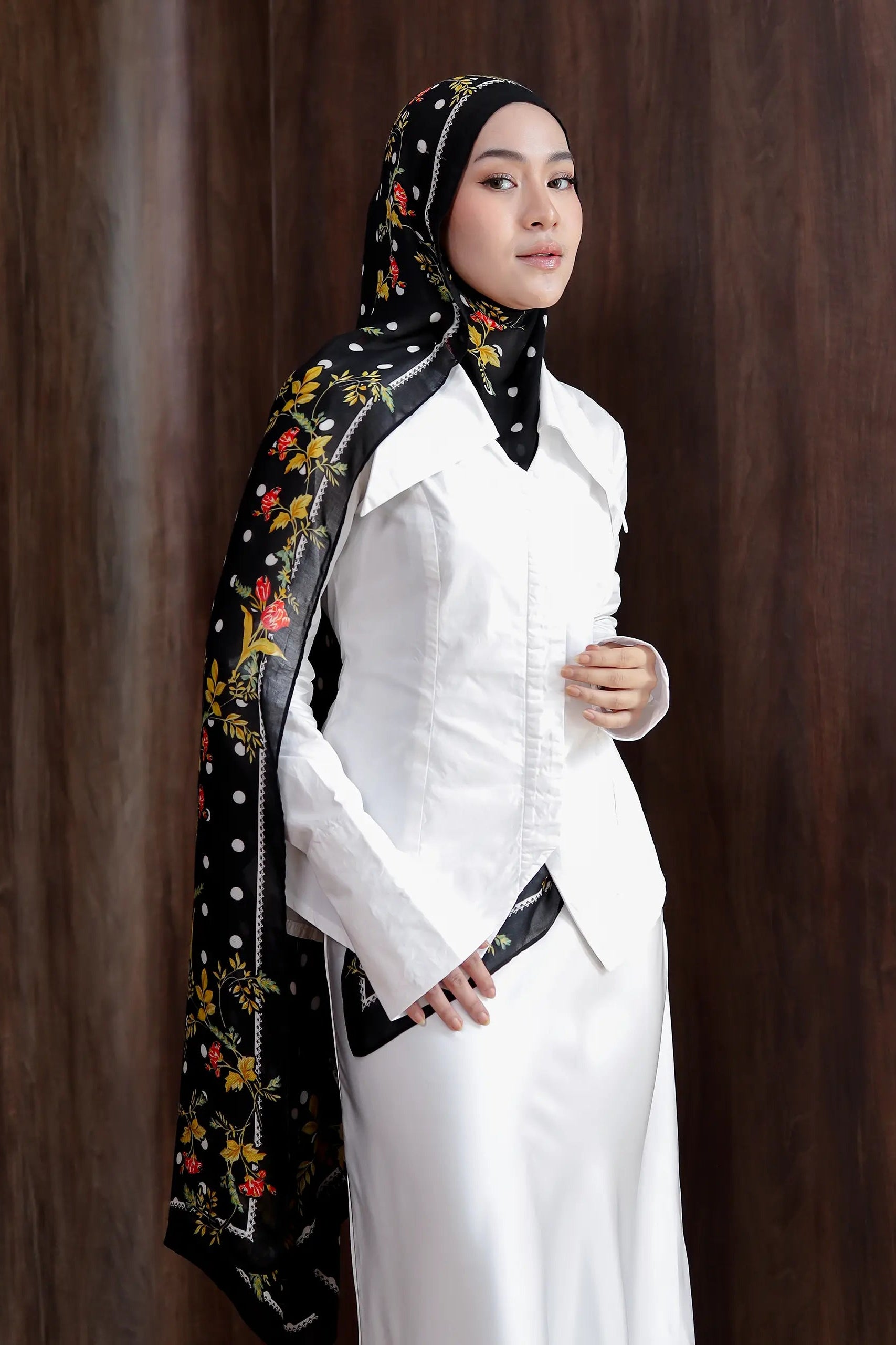 Woman wearing a white outfit with a black hijab featuring floral embroidery against a brown background