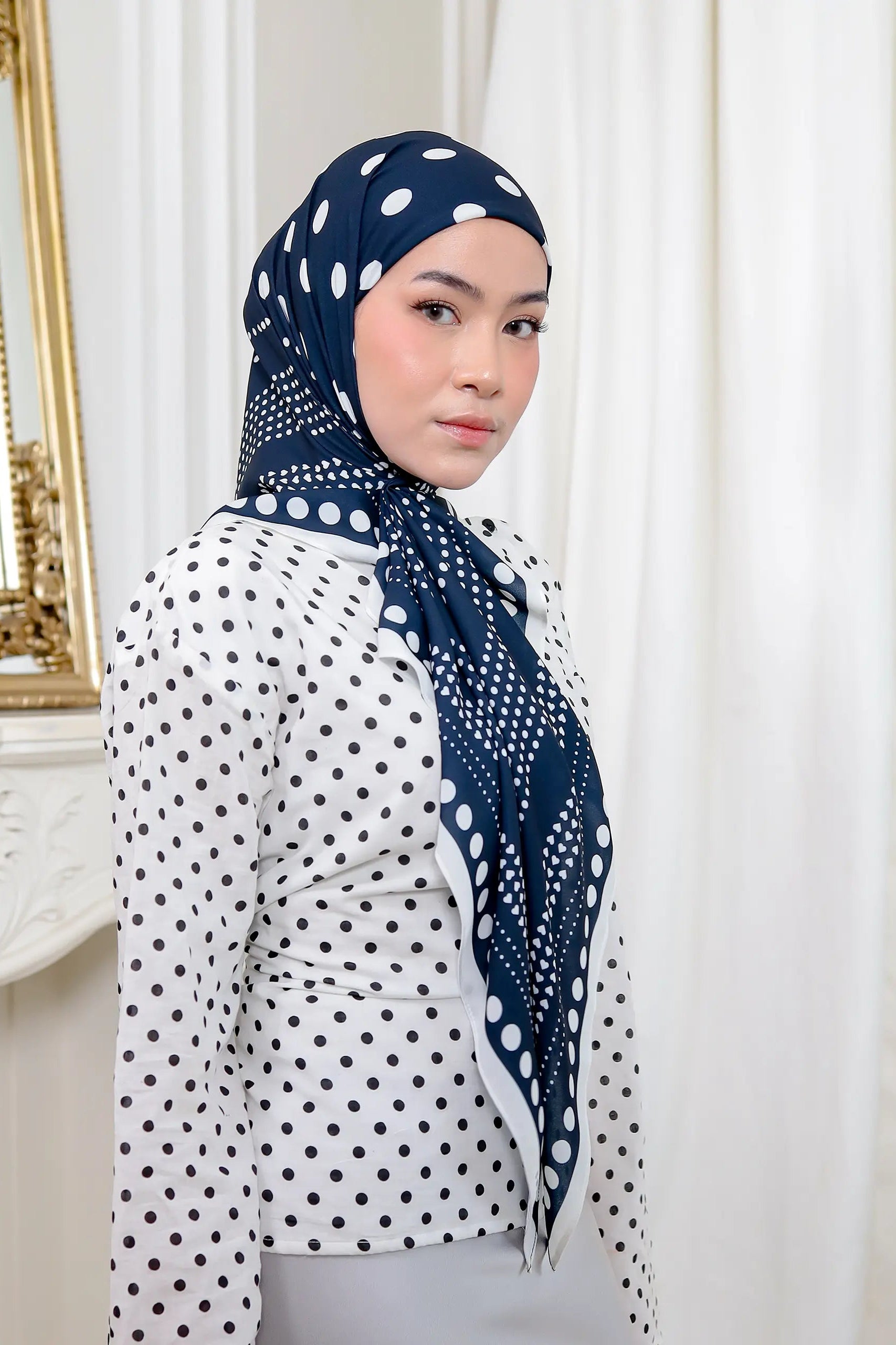 Woman wearing a navy blue hijab with white polka dots in a room with a mirror and decorative elements.