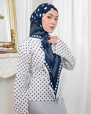 Woman wearing a white blouse with black polka dots and a navy hijab with white patterns, standing in a room with decorative elements.