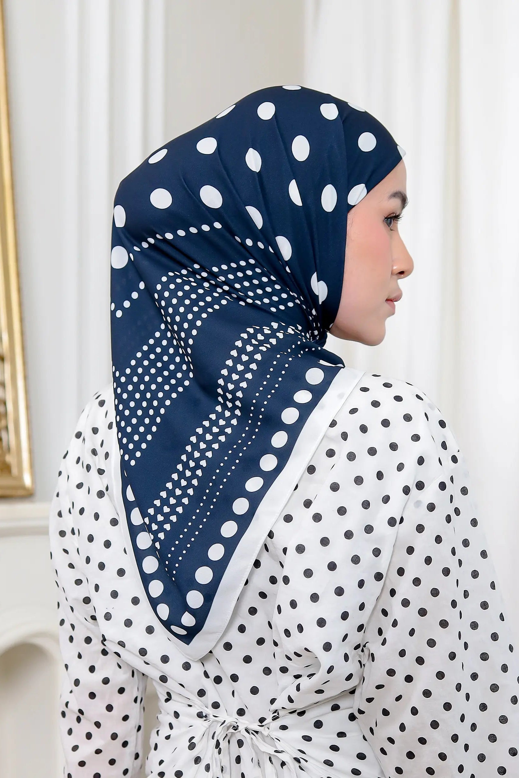 Woman wearing a navy hijab with white polka dots in a room with a mirror and white walls.