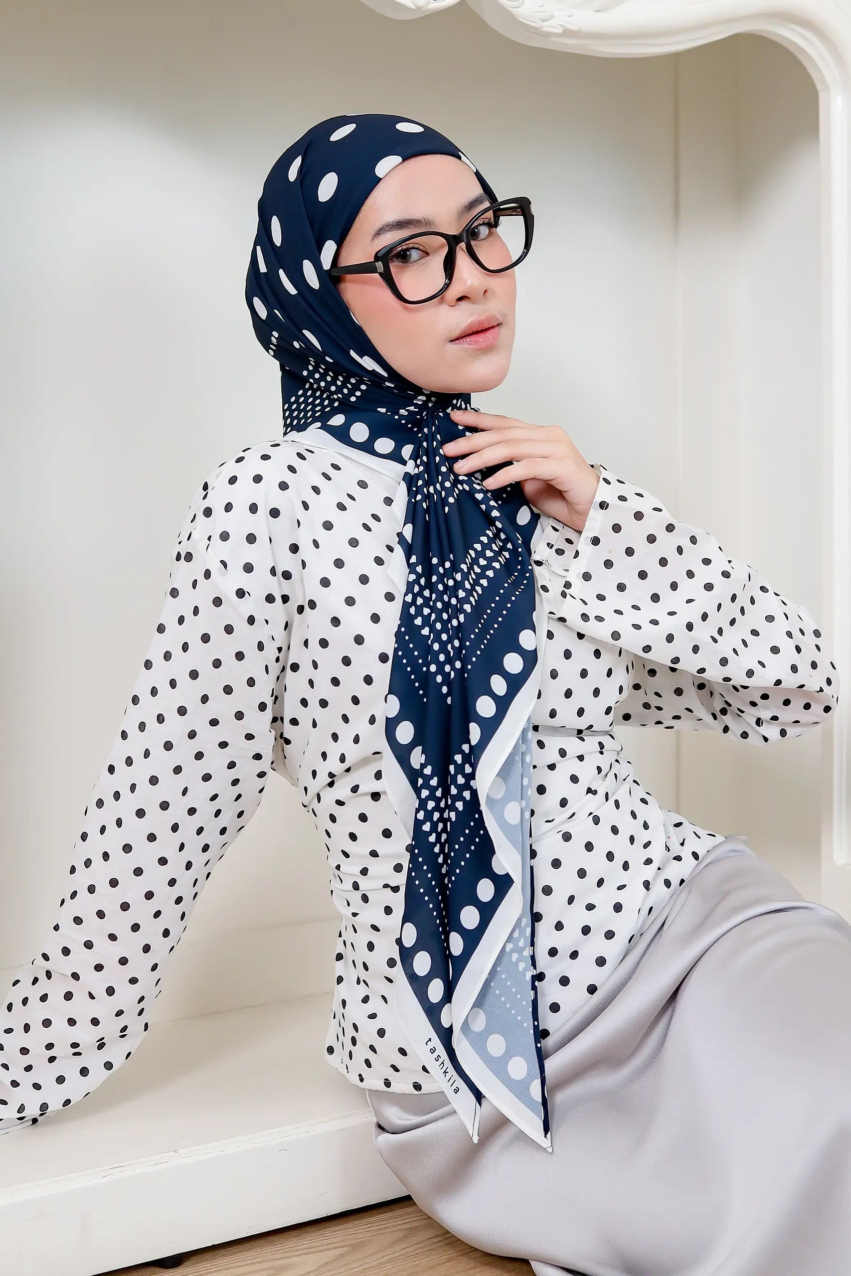 Woman wearing a polka dot headscarf and glasses in a white room.