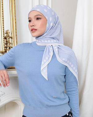 Woman wearing a light blue sweater and polka dot hijab in a decorative room.