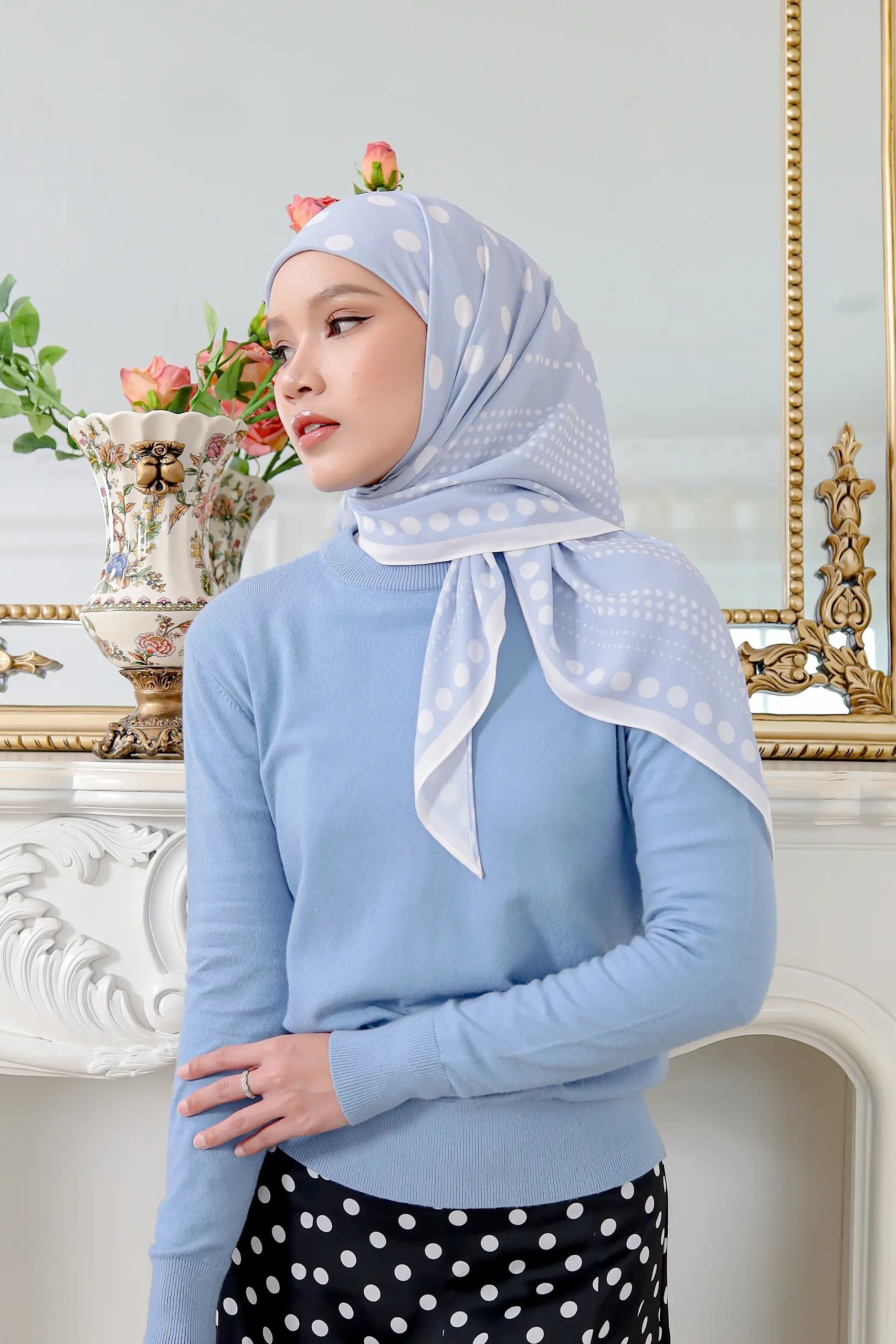Woman wearing a light blue hijab and sweater in a decorated room with flowers and a mirror.