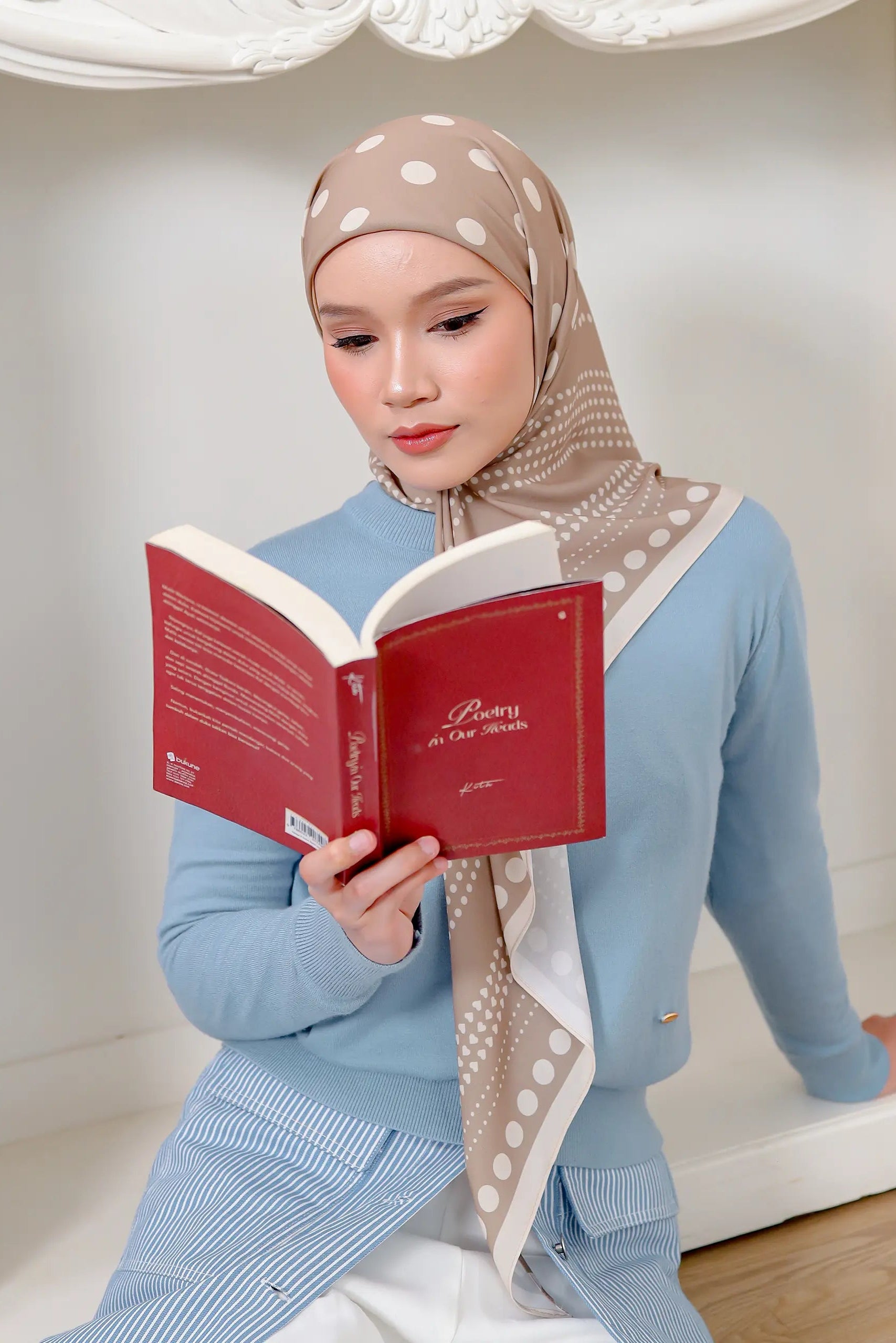 Woman reading a red book wearing a light blue outfit and patterned hijab.