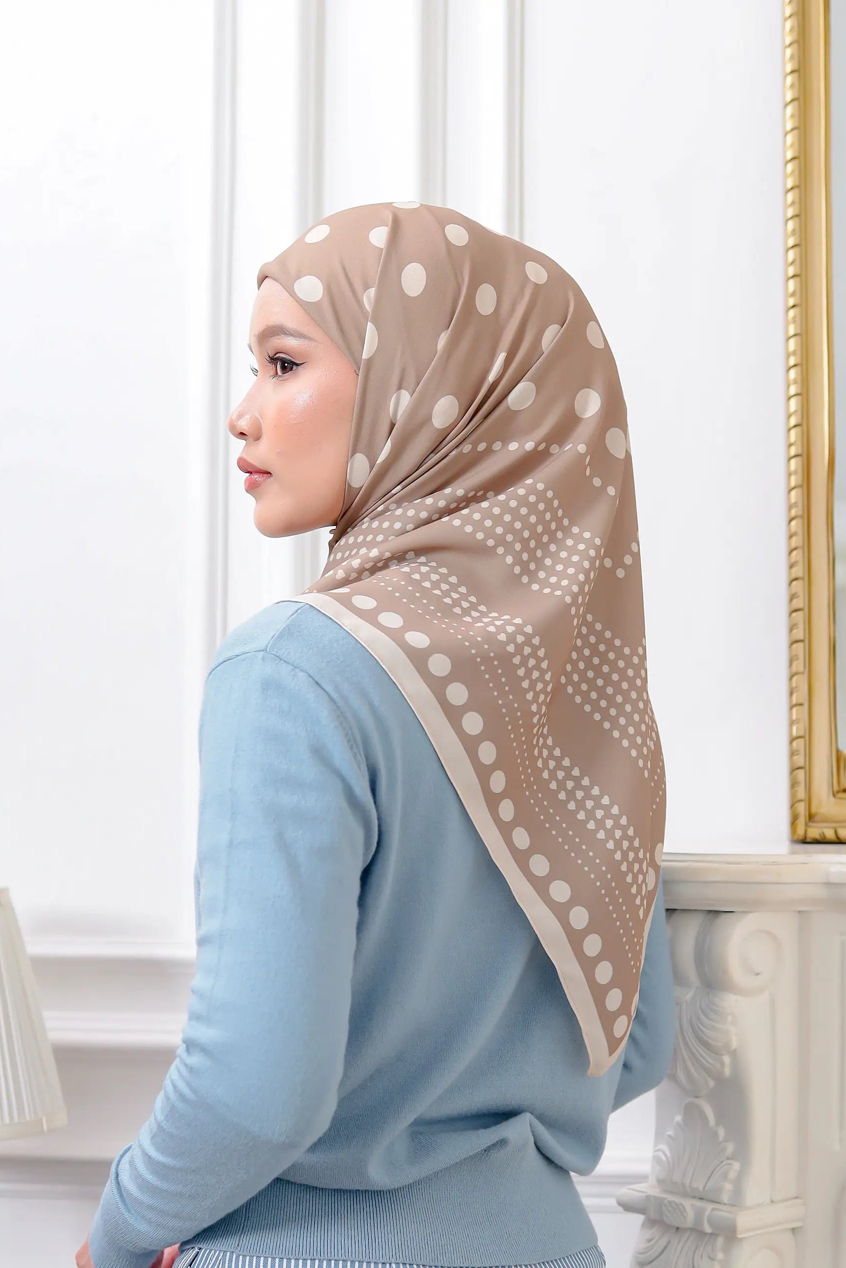 Woman wearing a beige hijab with white patterns in a room with a mirror and lamp.