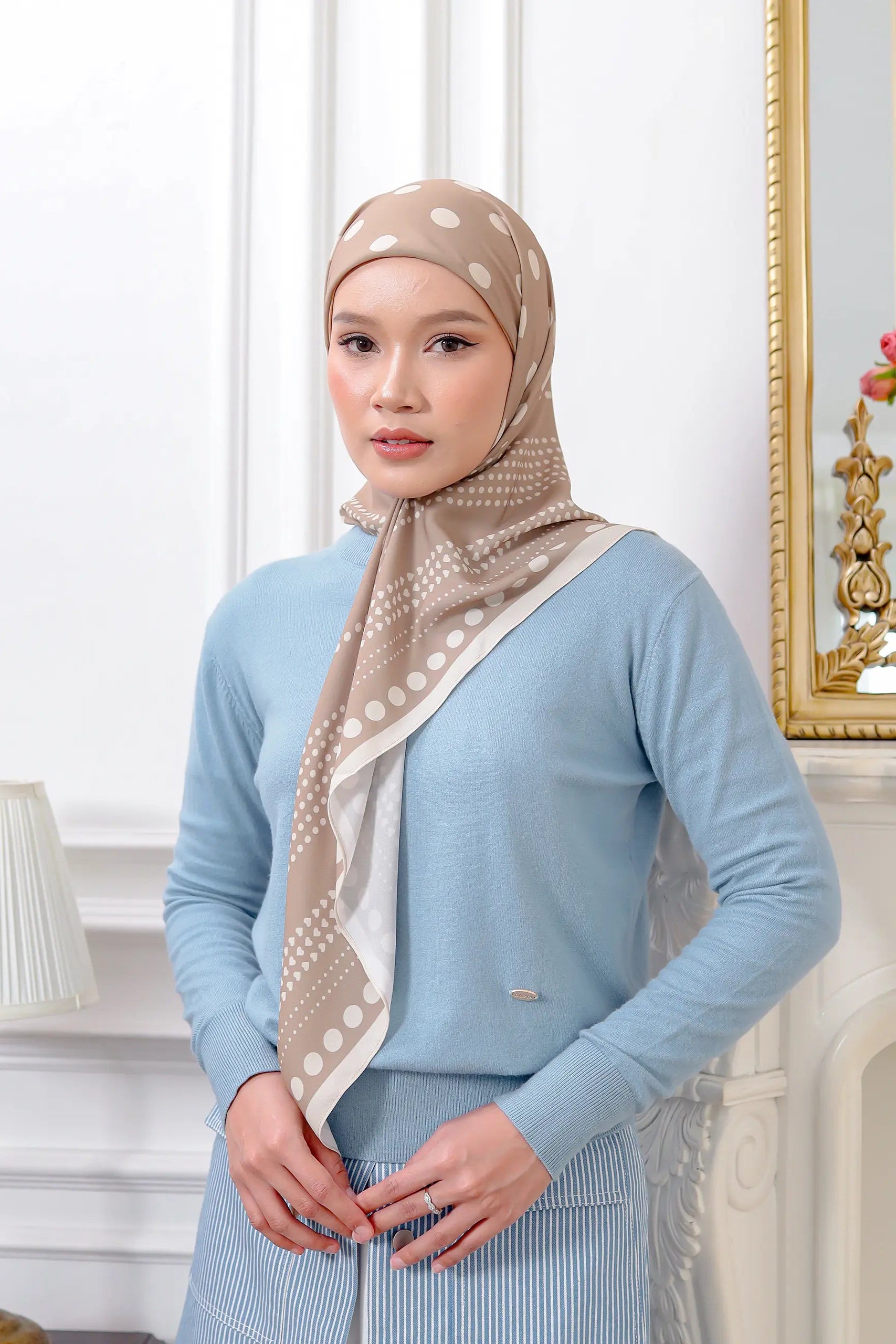 Woman wearing a light blue top and beige hijab with white patterns in an elegant room.