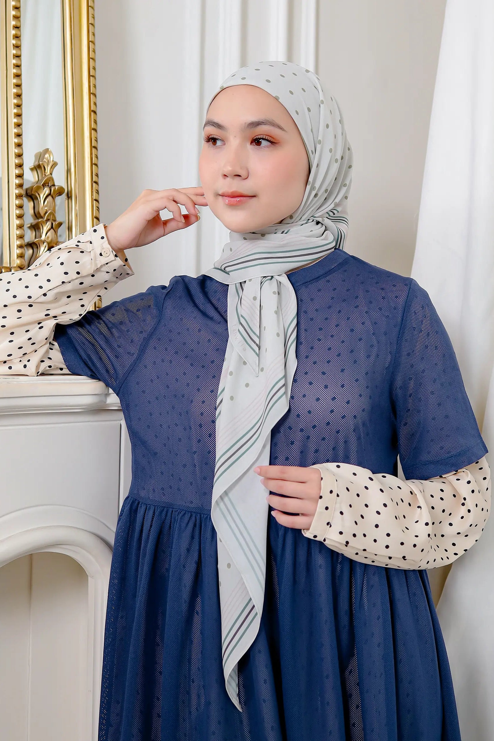 Woman wearing a blue dress with a white polka dot blouse and a gray hijab with white patterns, standing in a room with decorative elements.