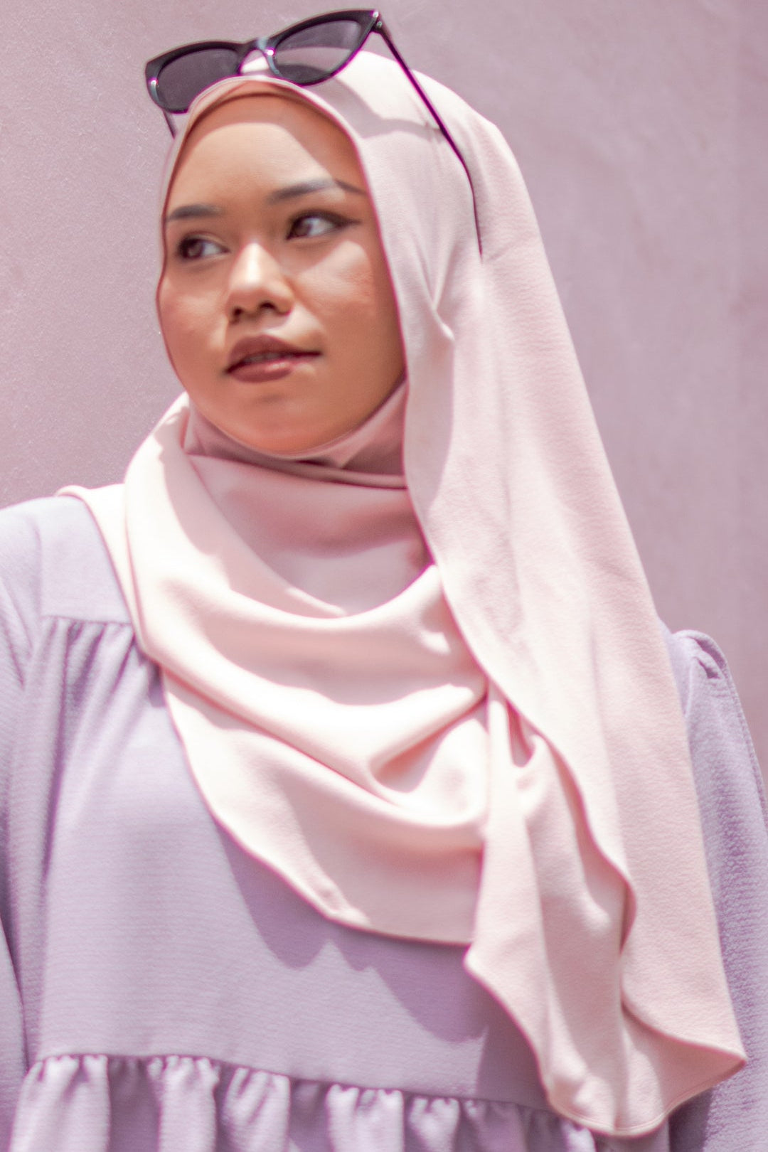 Woman wearing a light pink hijab and sunglasses on her head against a plain background