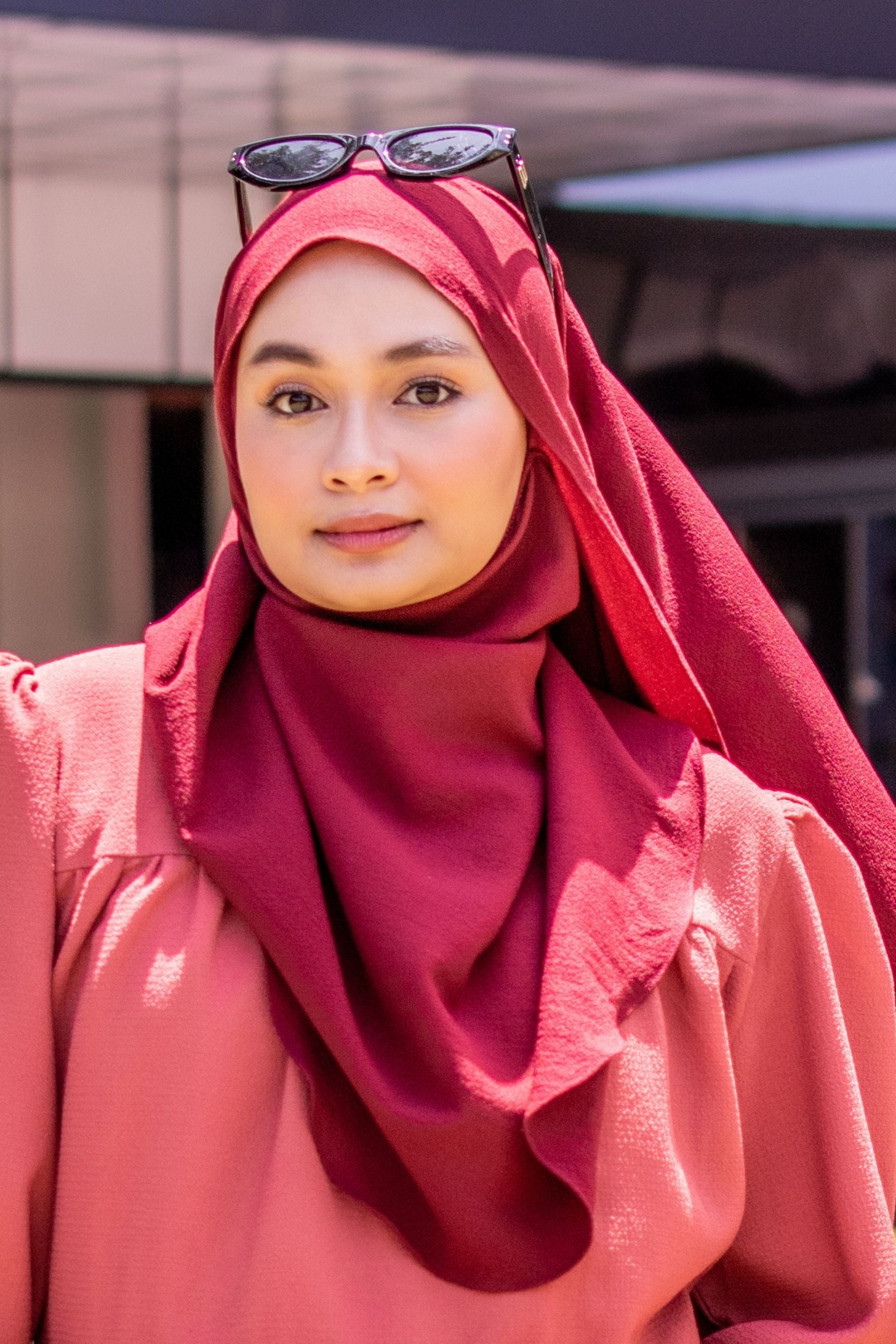 Woman wearing a red hijab with sunglasses on her head, standing outdoors.