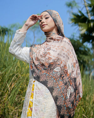Woman wearing a patterned hijab and dress in a natural setting with trees and blue sky.